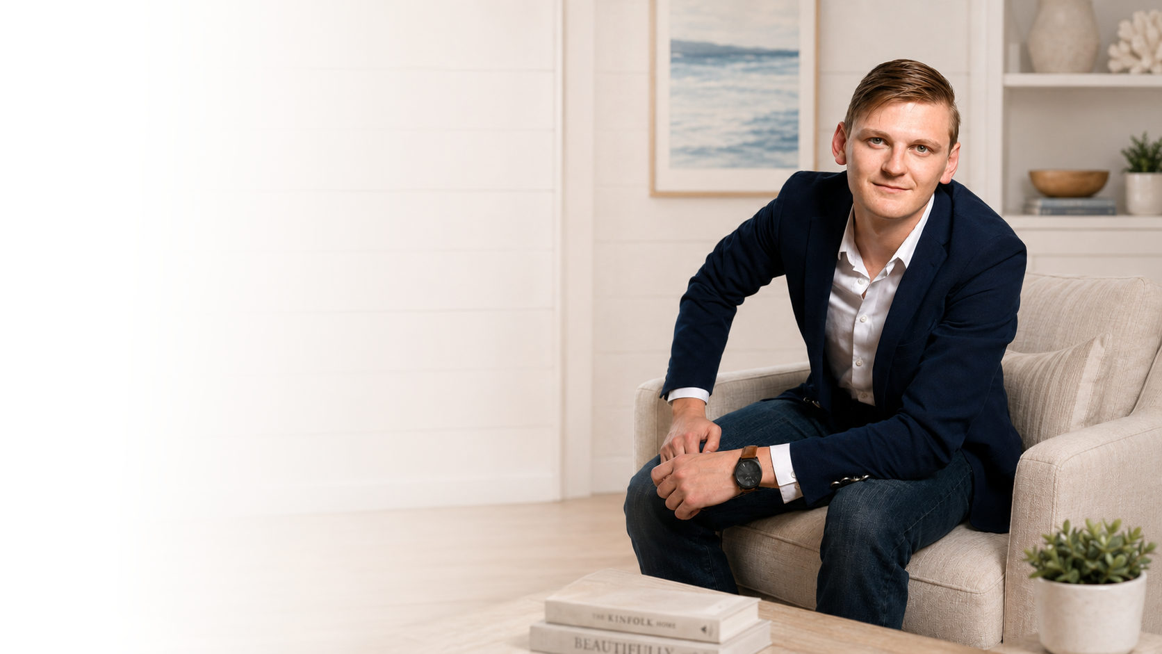 Real estate agent Devin Carver sitting on a beige sofa in a modern, minimalistic living room with a book on the table and artworks on the wall.