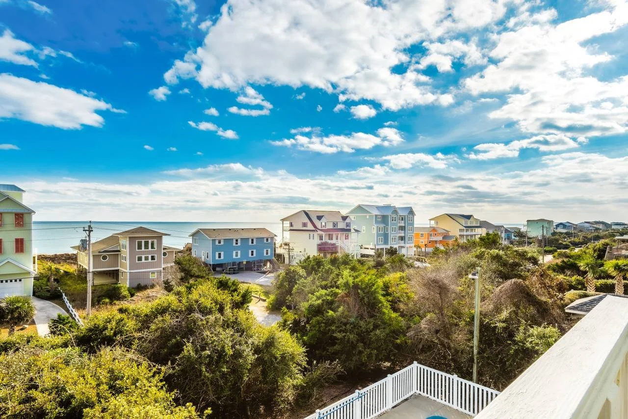 Colorful houses on a coastal neighborhood with trees and a view of the ocean under a partly cloudy sky.