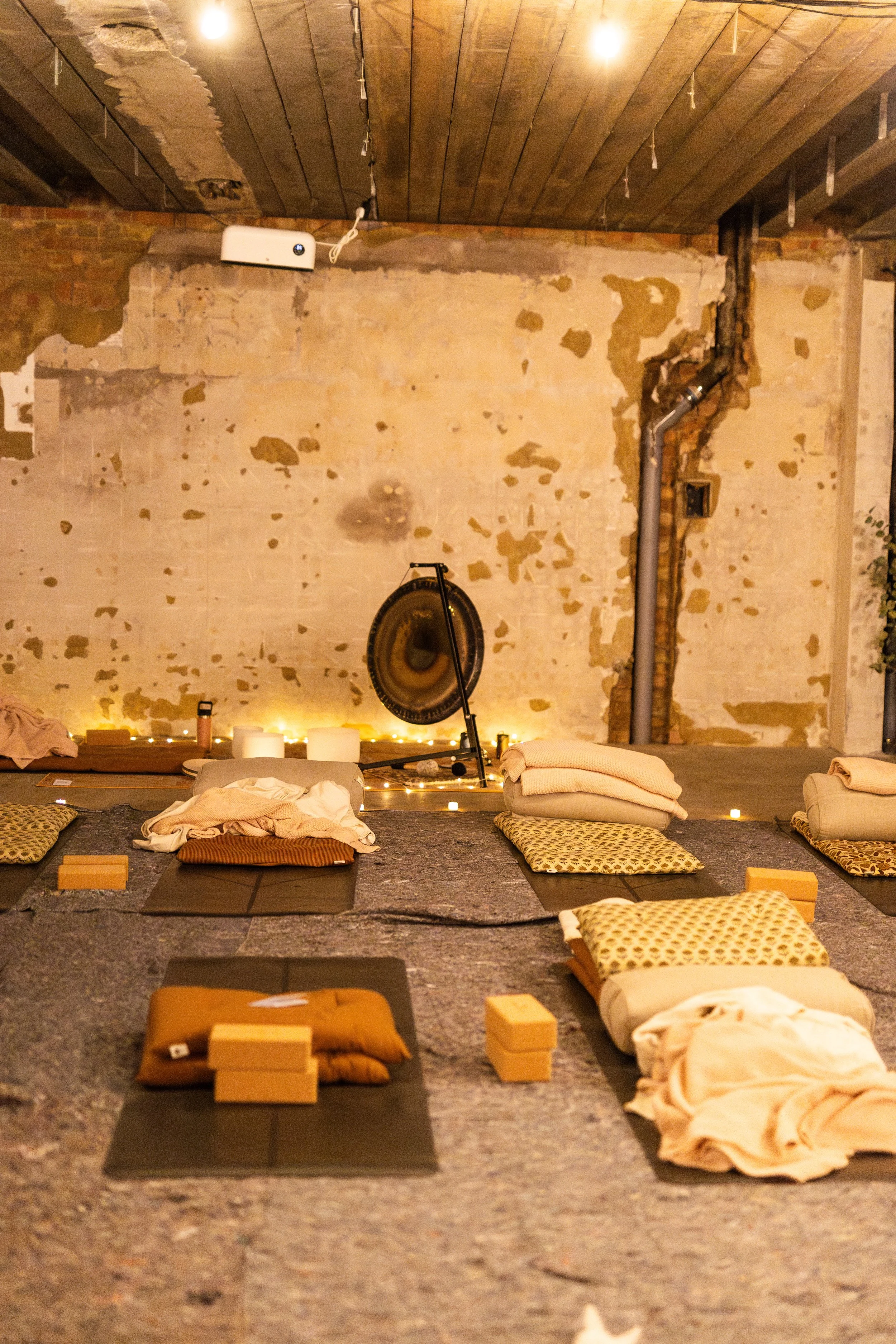 A yoga or meditation space with mats and cushions arranged on a textured stone or concrete floor. There are lit candles along the back wall, which features an exposed brick and plaster surface with some peeling paint. The room has a rustic, industrial vibe with a wooden ceiling and a large metal gong in the background. A projection speaker and a pipe are visible on the wall.