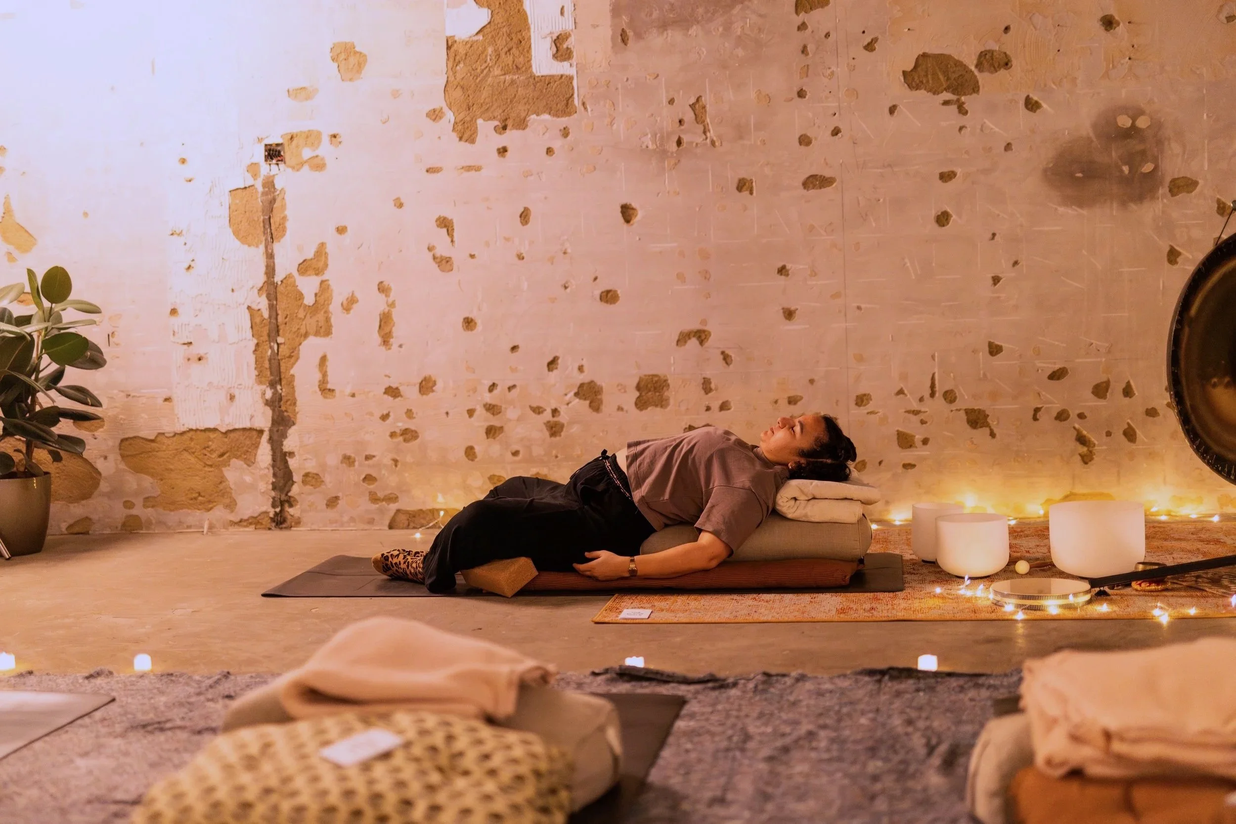 A woman lying on a yoga mat in a wellness or meditation room with a distressed wall, surrounded by candles and plants, appearing relaxed and peaceful.