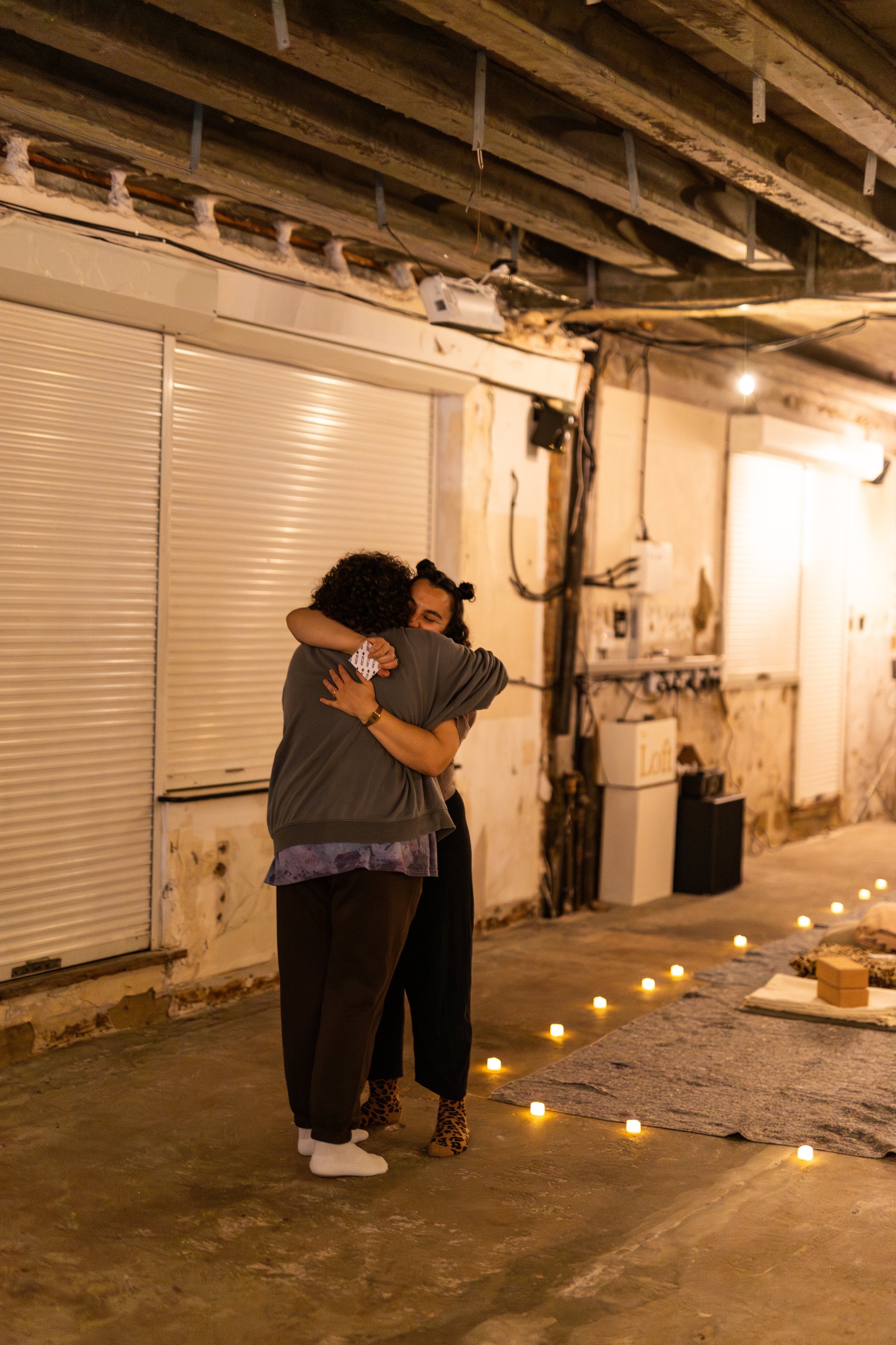 Two people hugging in a warmly lit industrial-style indoor space with candles on the floor, closed metal shutters, and exposed pipes and wiring on the ceiling.