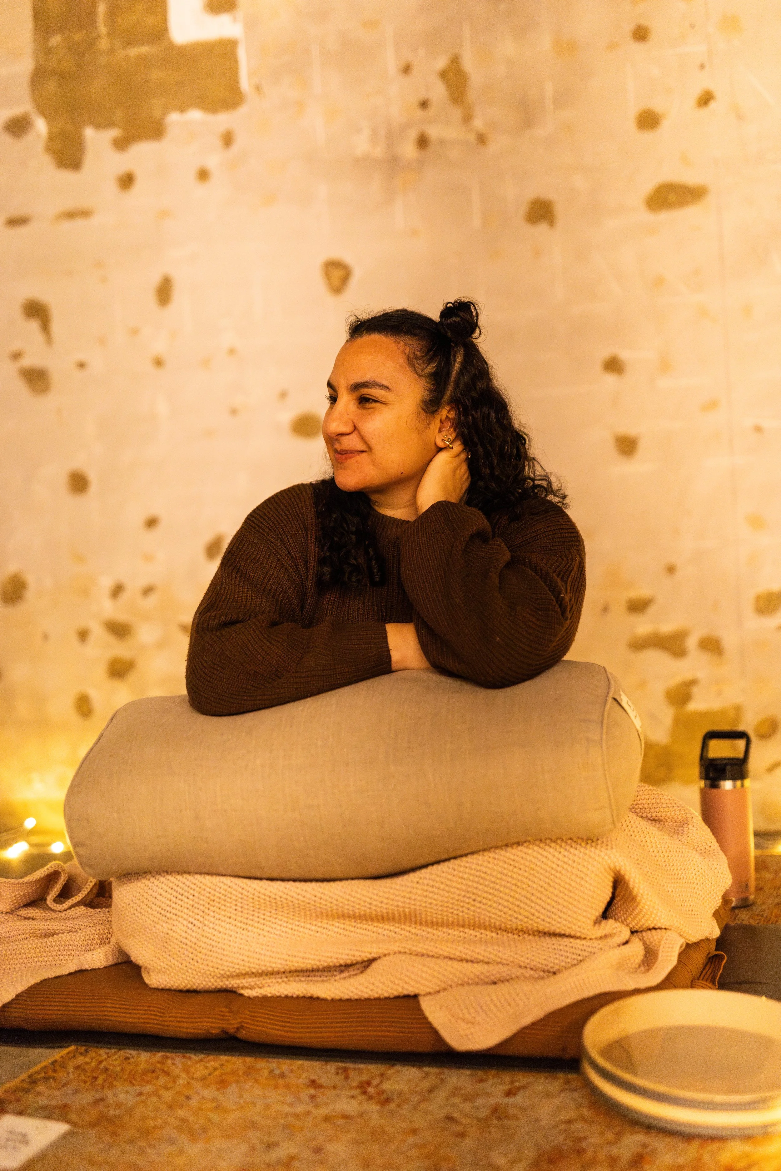 A woman with curly dark hair, wearing a brown sweater, is sitting with her arm resting on a cushion in an indoor setting with a distressed concrete wall in the background.
