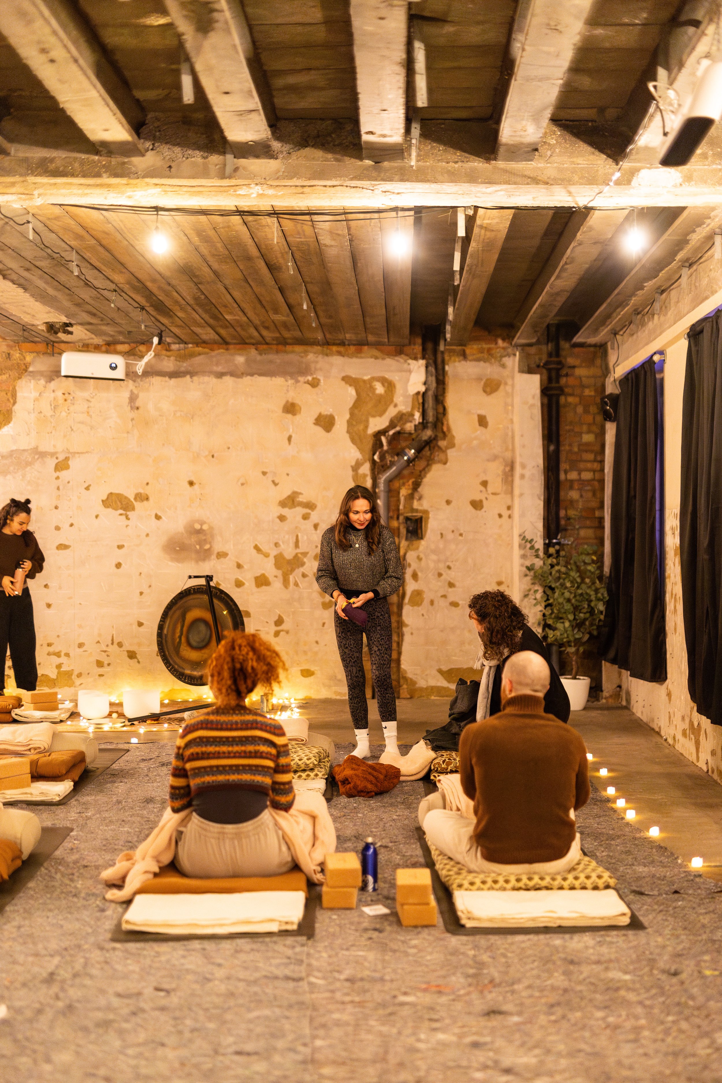 People participating in a yoga or meditation session in a loft-style room with exposed brick and wooden beams, seated on cushions on the floor facing a woman standing at the front.