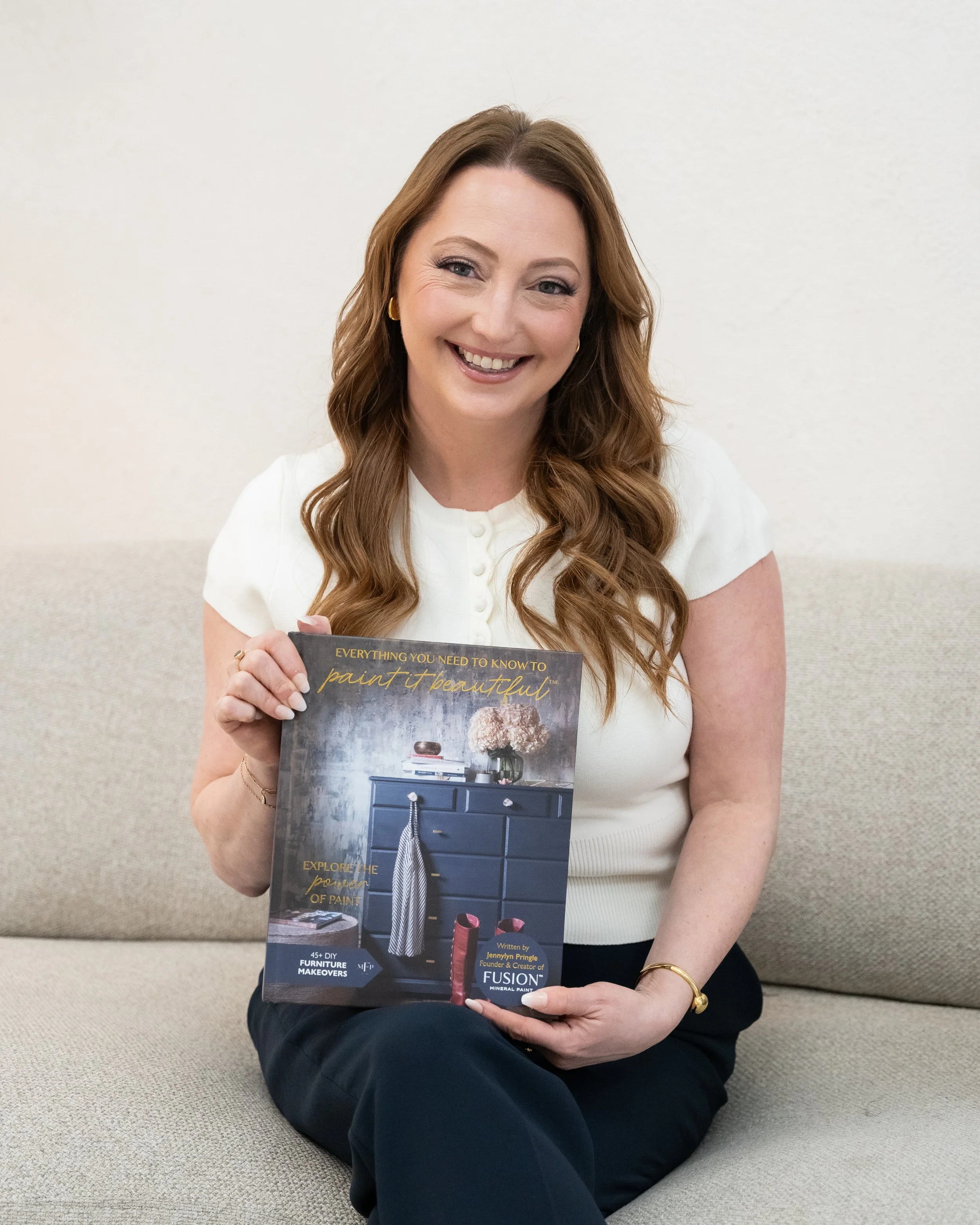 A woman with long wavy hair, wearing a white top, sitting on a beige sofa against a plain light-colored wall, smiling, holding a magazine titled 'Everything You Need to Know to Paint Beautiful'.