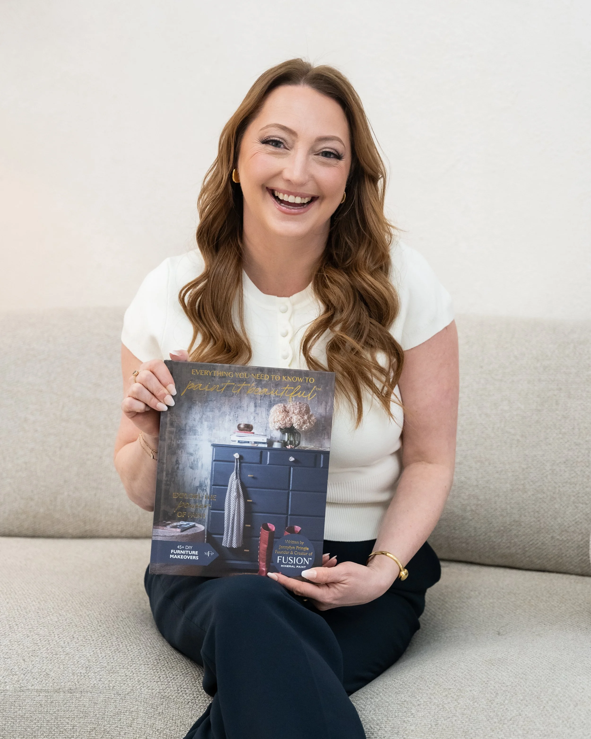 A woman with long, wavy auburn hair and gold earrings, smiling, sitting on a beige carpeted floor holding a magazine about interior paint and furniture.