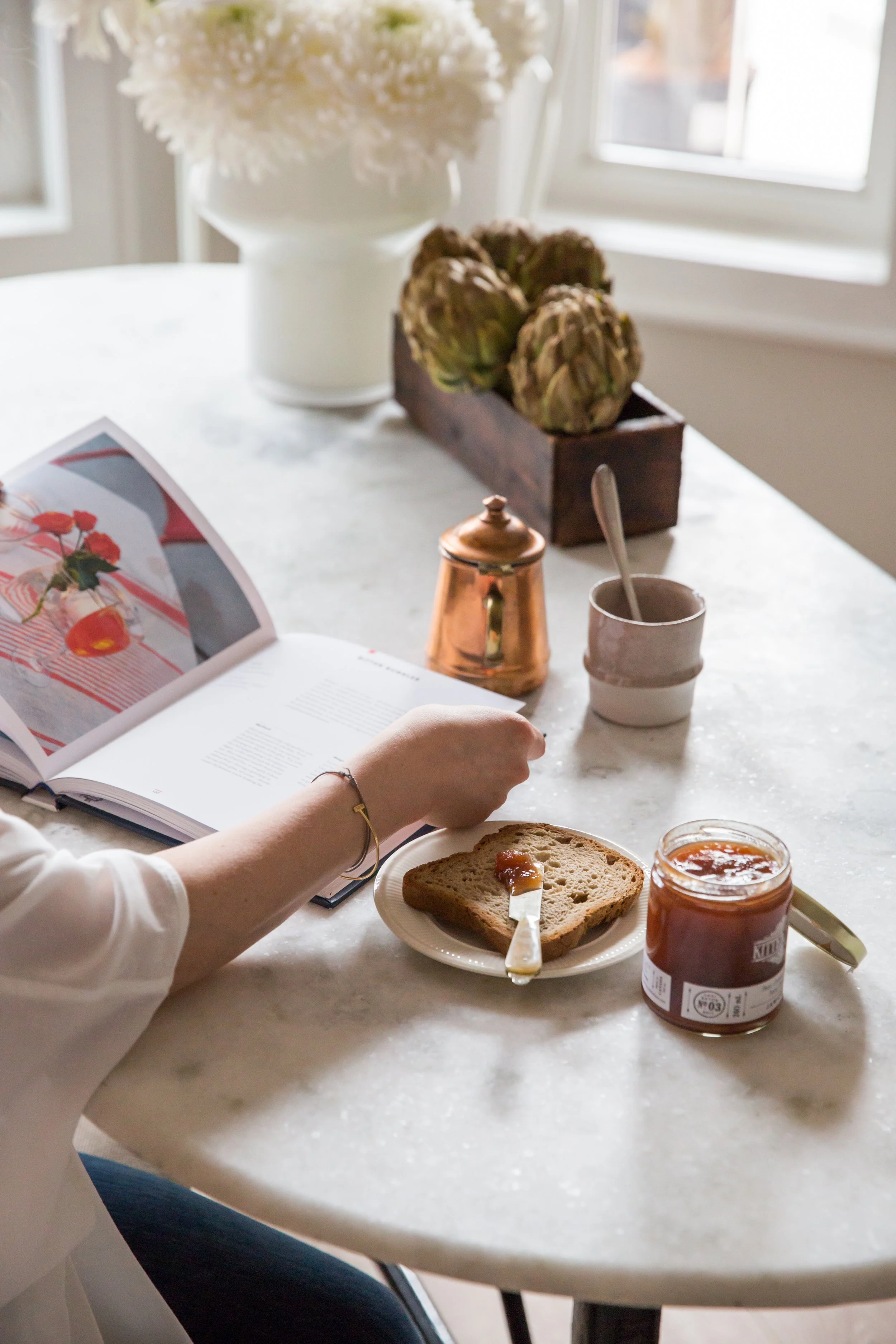 A breakfast scene on a white marble table, featuring a person spreading jam on toast, a jar of jam, a small butter knife, a white cup, a copper teapot, a magazine, a bowl of white flowers, artichokes in a wooden box, and a potted plant with greenery near a window.