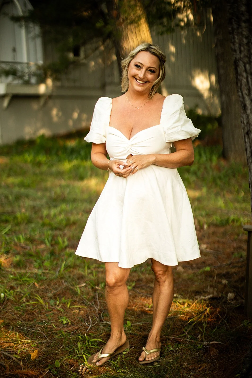 A woman in a white dress standing outdoors in a yard with trees and grass, smiling at the camera.