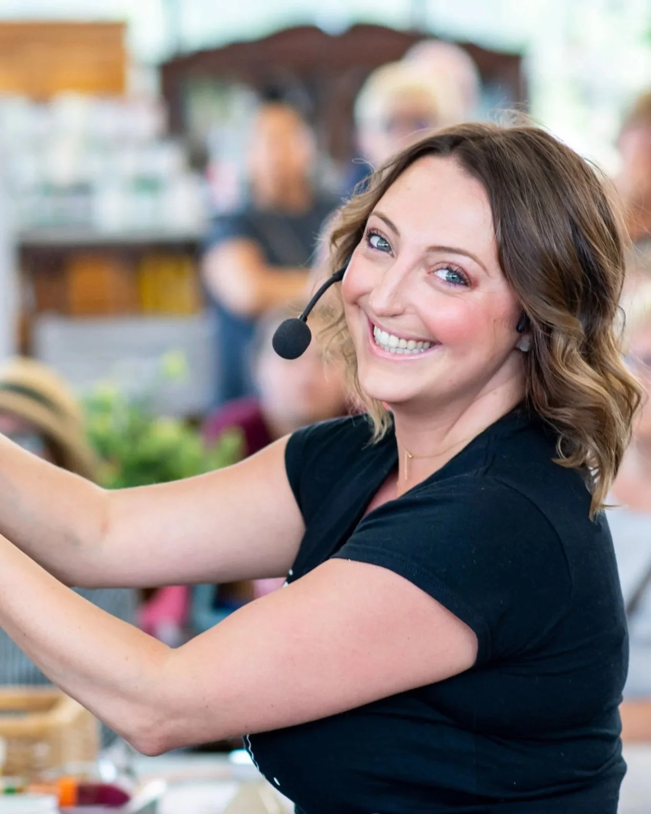 A woman with wavy brown hair, smiling, wearing a headset, taking a selfie or video call, in a busy indoor setting.