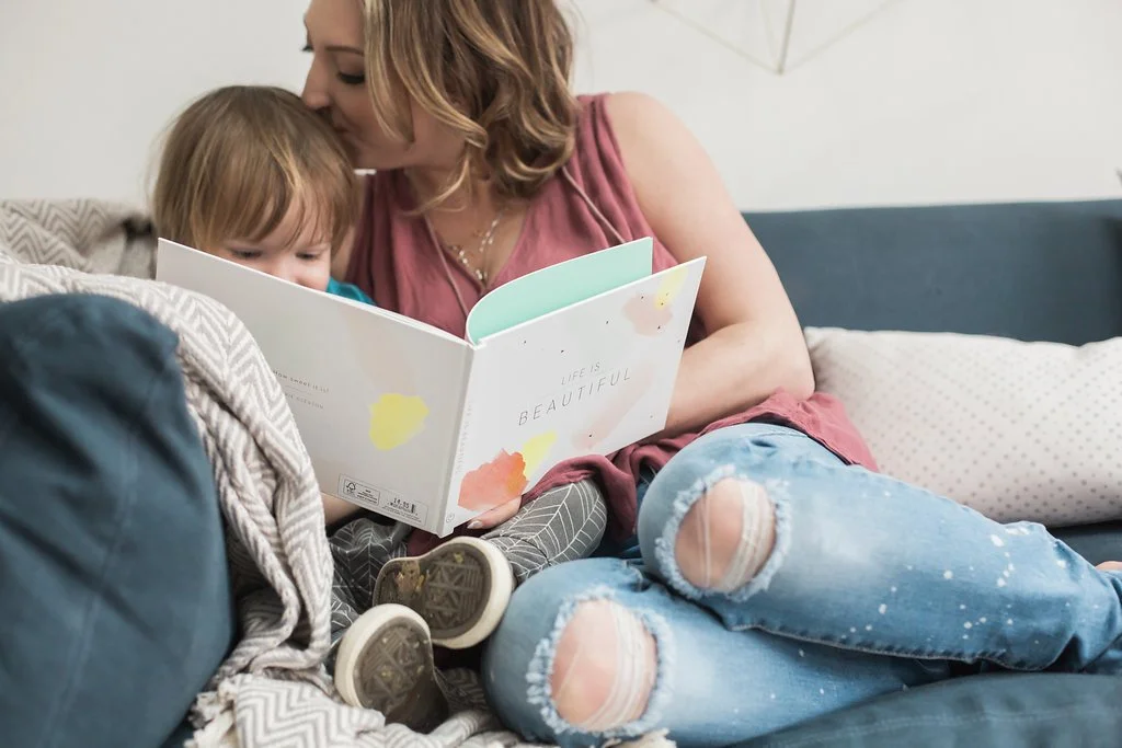 A woman with shoulder-length hair and a pink sleeveless top reads a colorful children's book to a young child with short hair, sitting on a couch with pillows, both leaning in and sharing the moment.