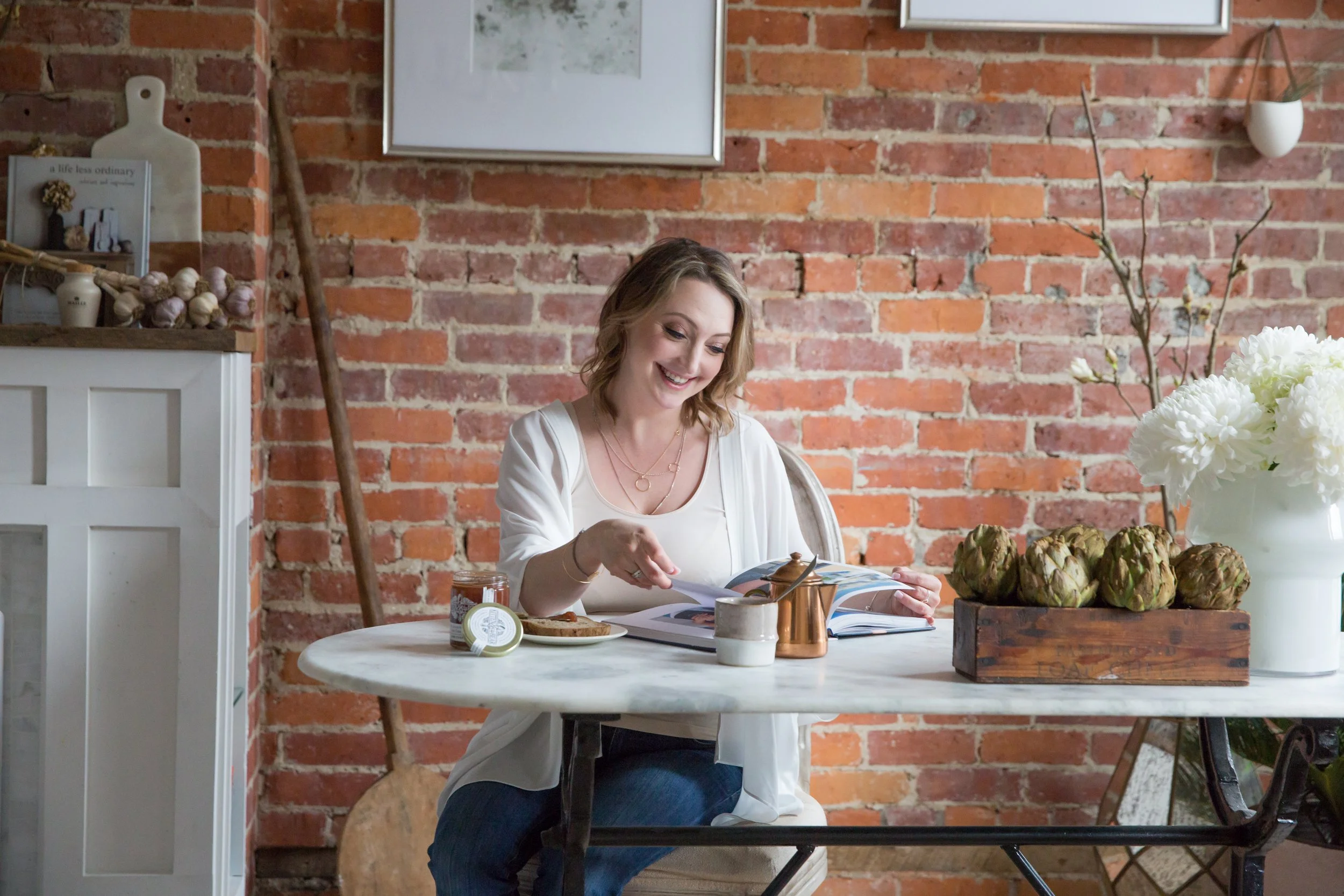 A woman with shoulder-length light brown hair is sitting at a round white table reading a magazine. The table has a jar of jam, a small bowl, a cup, a copper pitcher, and a wooden tray with artichokes. The background features a red brick wall decorated with framed artwork, a white ceramic vase holding branches, and various kitchen or decorative items.