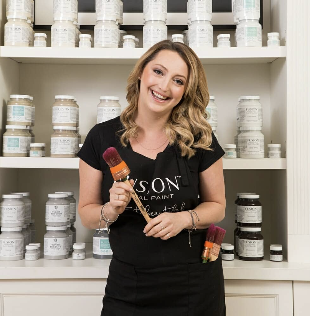 A smiling woman with wavy blonde hair holding makeup brushes standing in front of shelves filled with jars of Fusion mineral paint.