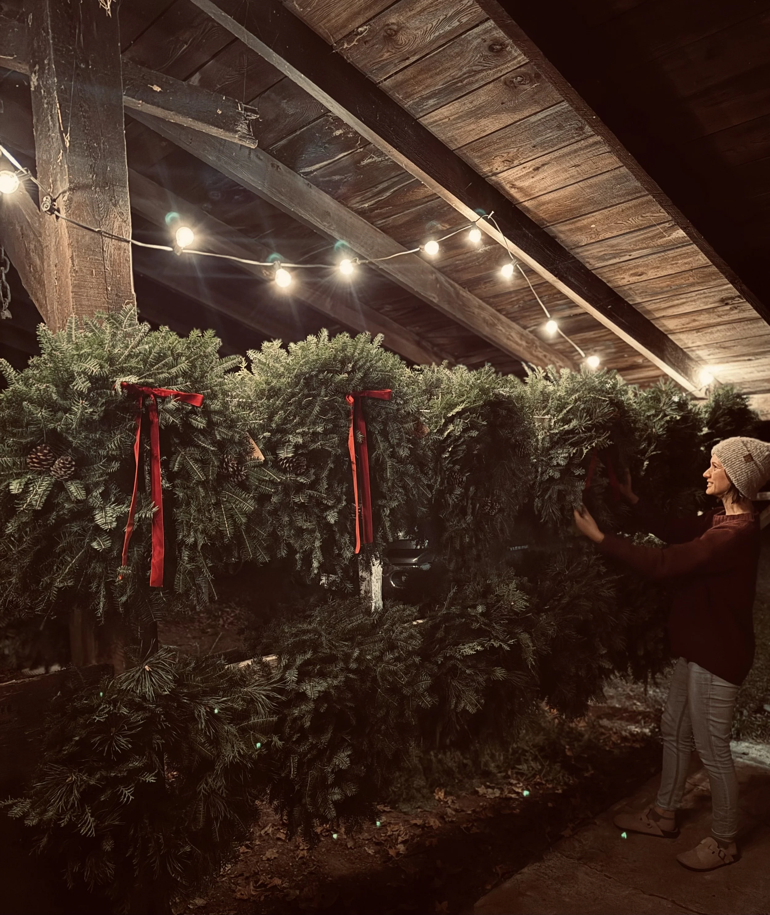 A woman wearing a gray knit hat and glasses stands next to a display of Christmas trees decorated with red ribbons. The setting is outdoors under a wooden porch ceiling with string lights.