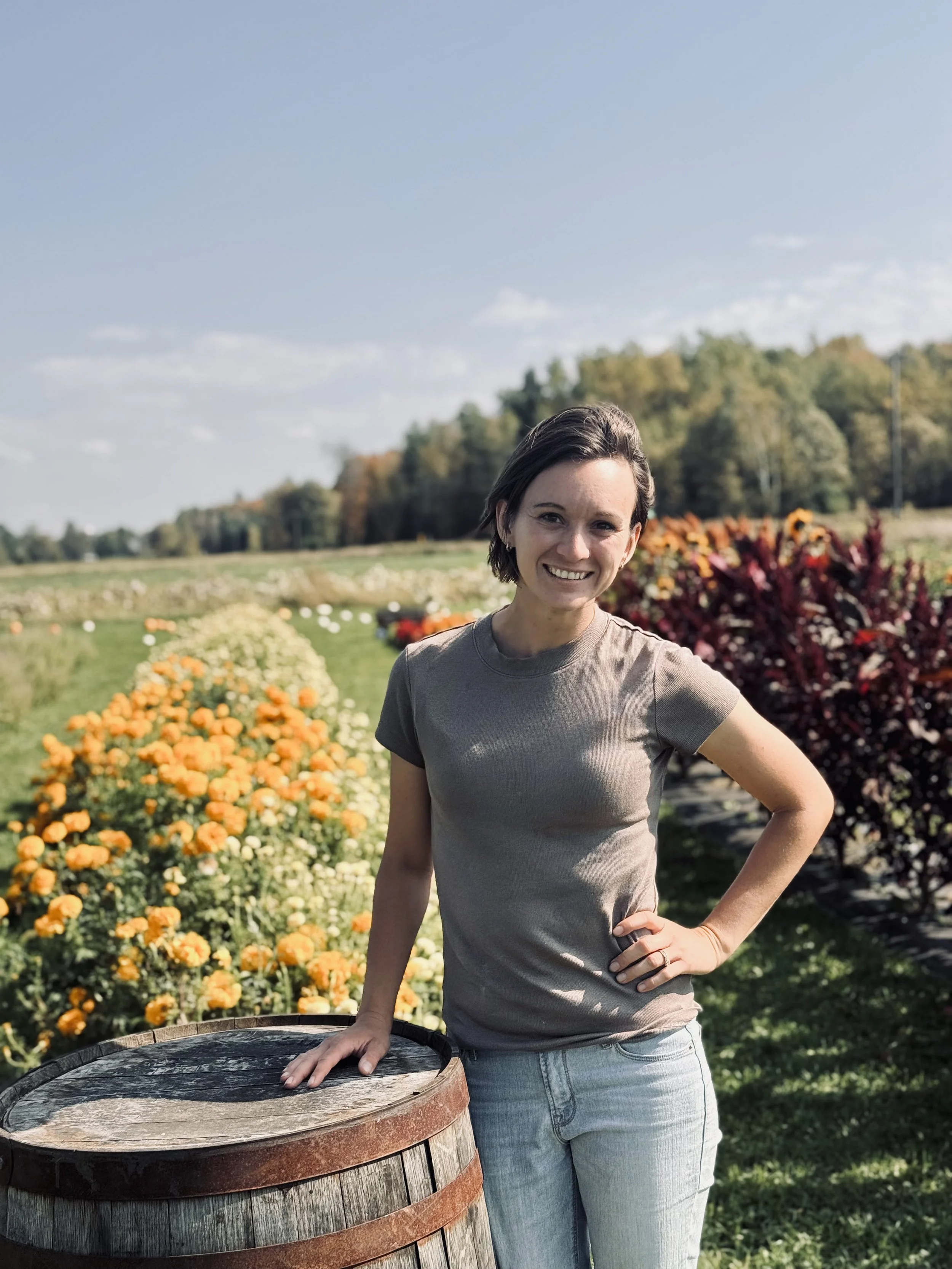 A woman standing outdoors in a rural setting with trees and flower beds in the background, smiling at the camera, wearing a beige t-shirt and light blue jeans, resting her hand on a wooden barrel.