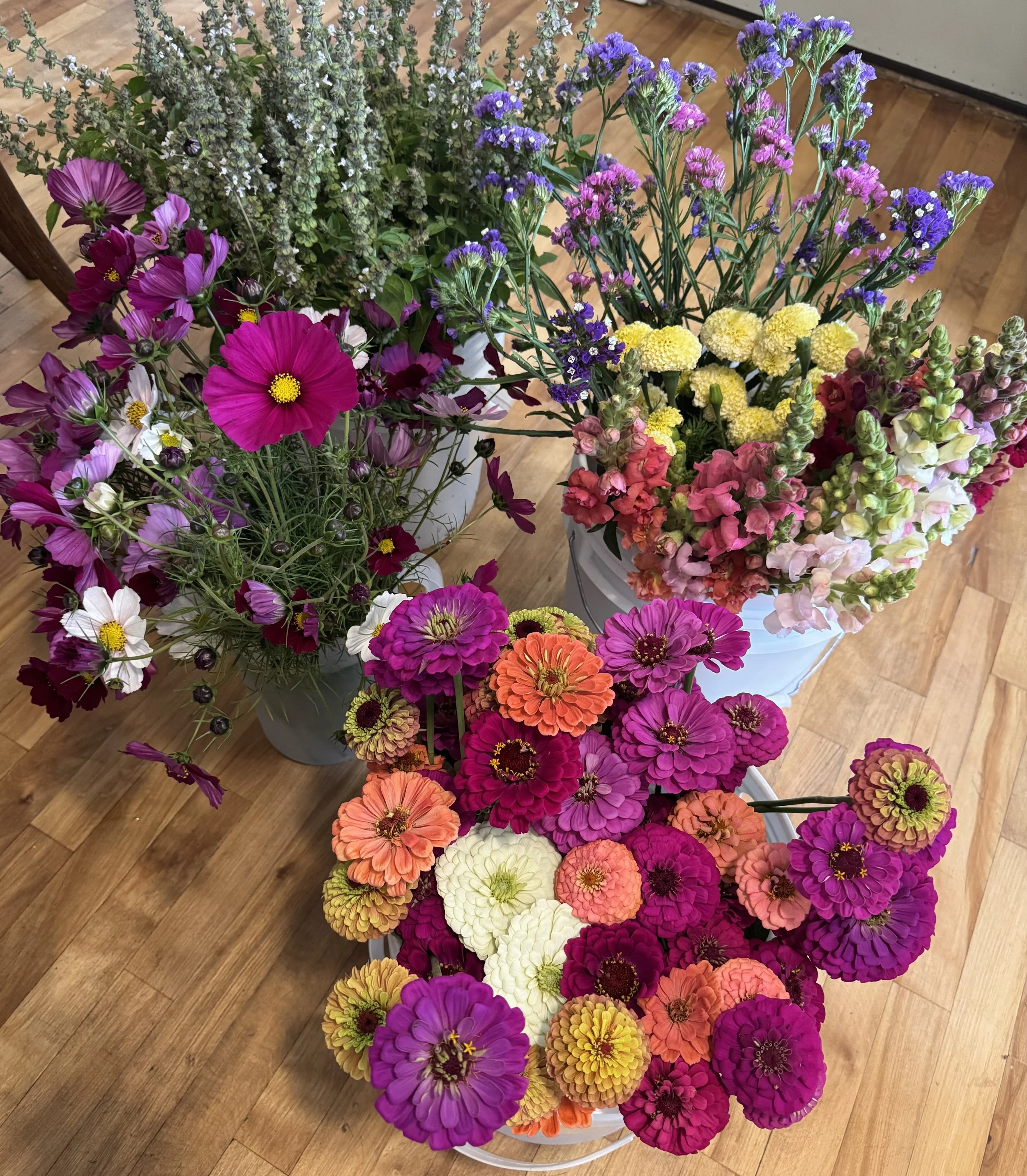 Colorful mixed flower bouquet including pink, purple, orange, yellow, and white blooms arranged in multiple containers on a wooden floor.