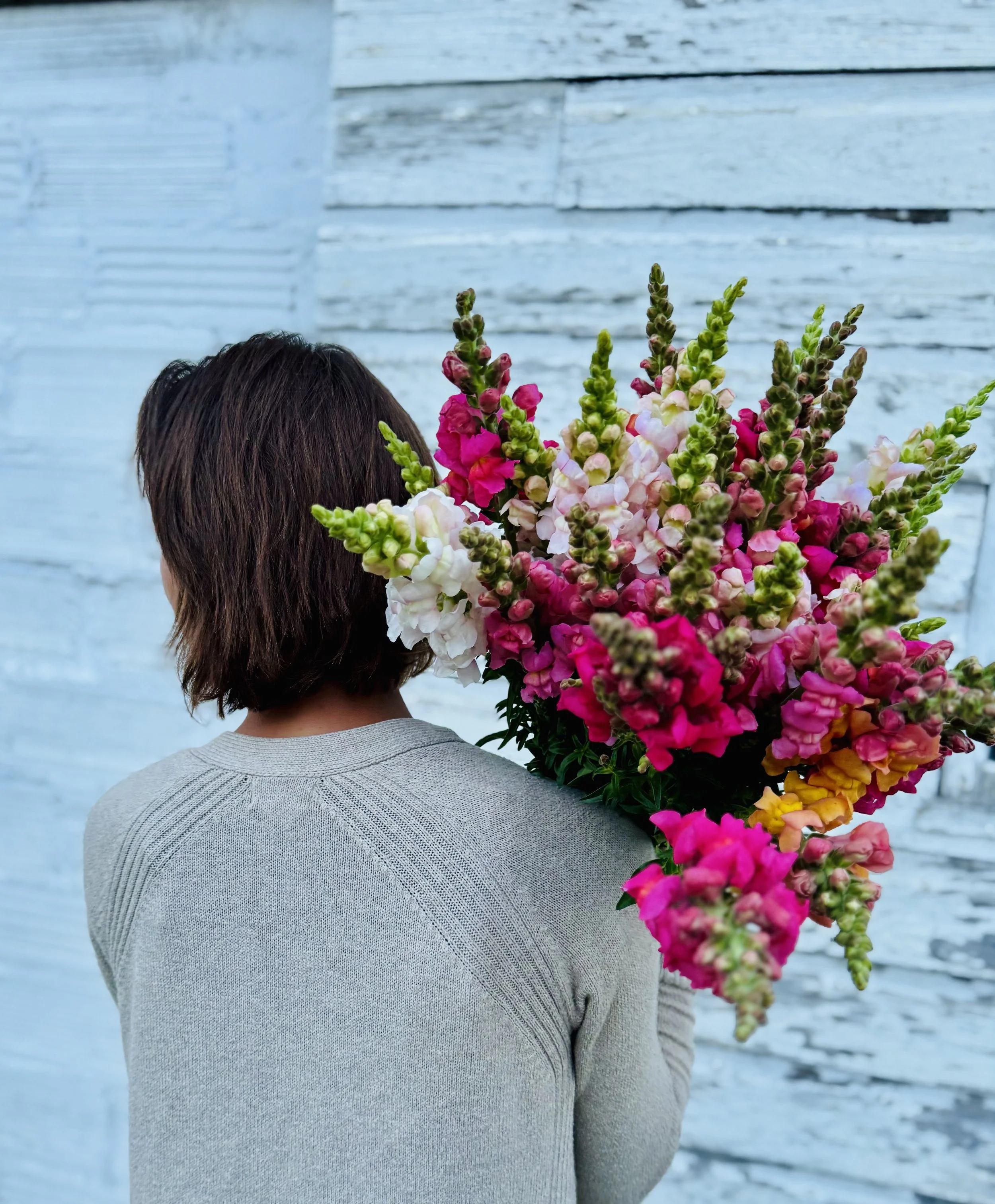 Person holding a large bouquet of colorful flowers, standing in front of a light blue wooden wall.