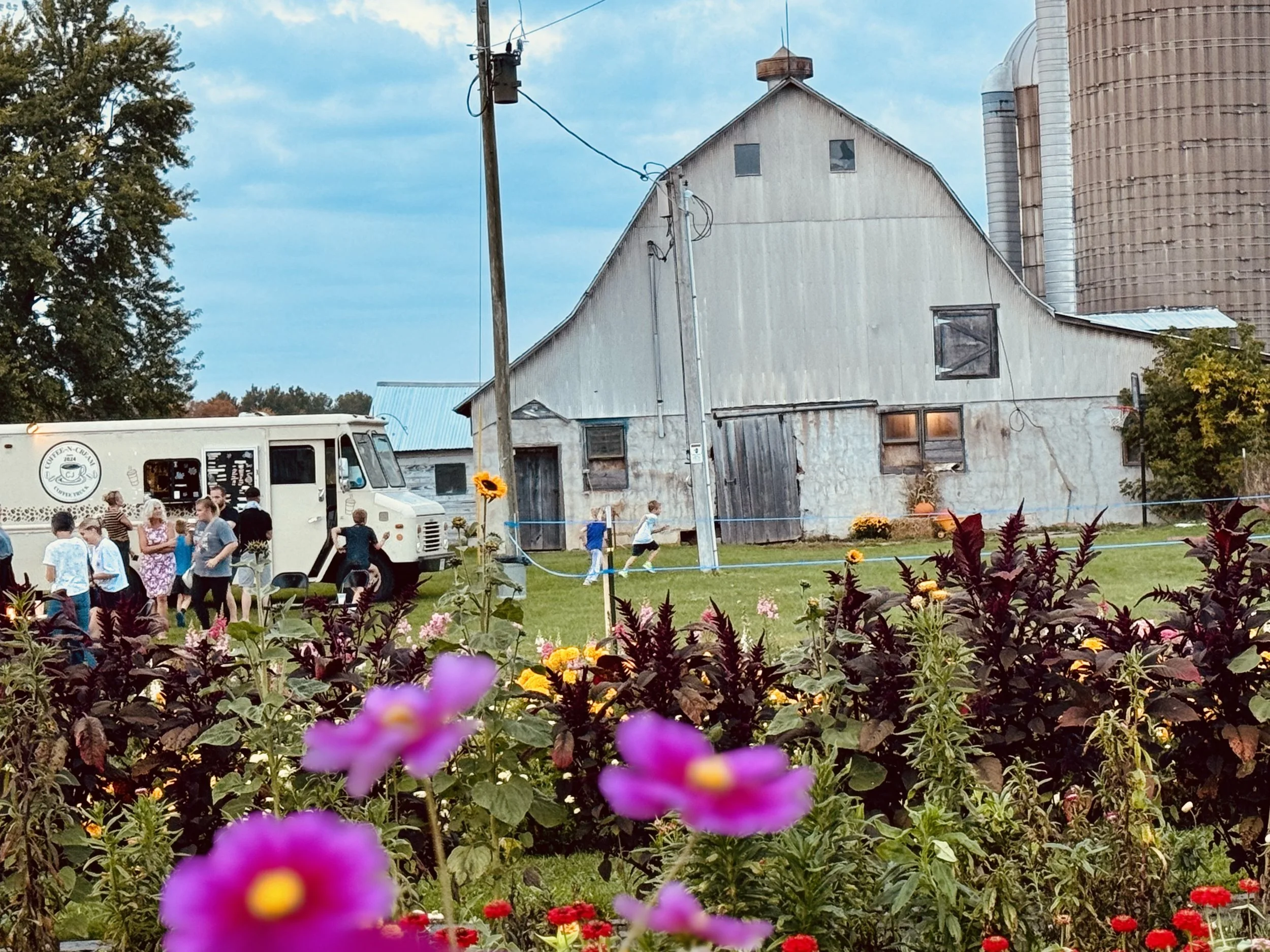A rural scene with a crowd gathered near a food truck, surrounded by colorful flowers in the foreground and an old barn with grain silos in the background, under a partly cloudy sky.