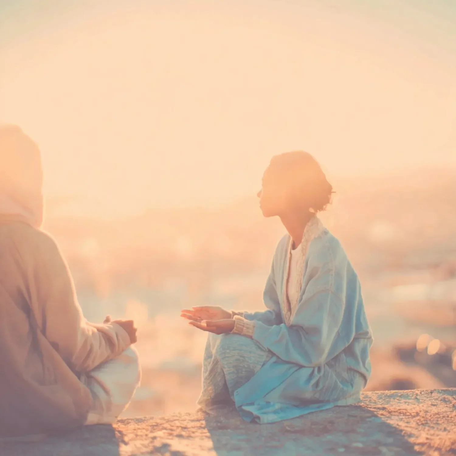 A man and woman sitting by the beach, holding hands, with the woman in a light blue dress and the man in beige, during sunset.
