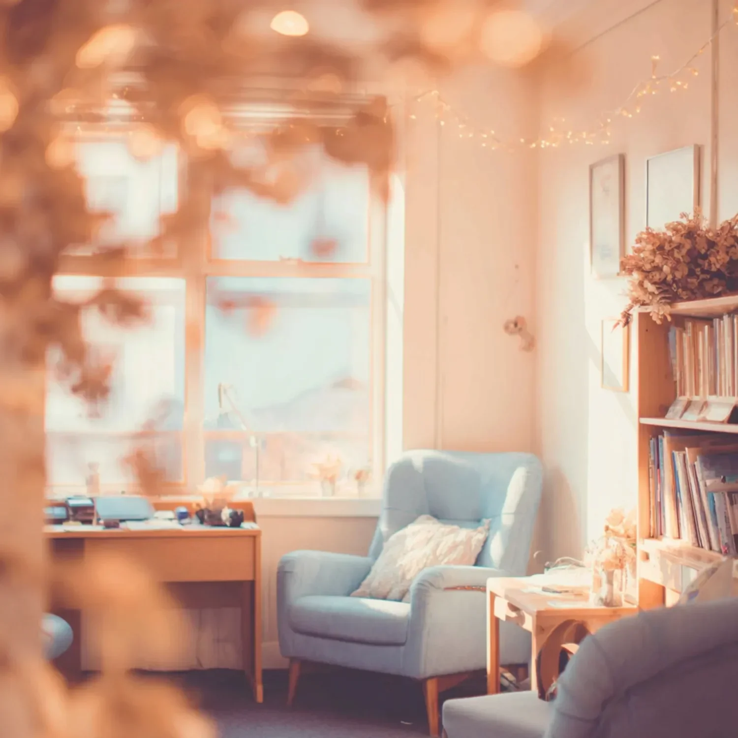 A cozy living room with a large window, a blue armchair with a floral pillow, a wooden side table, a bookshelf with books, and string lights hanging on the wall, illuminated by sunlight.