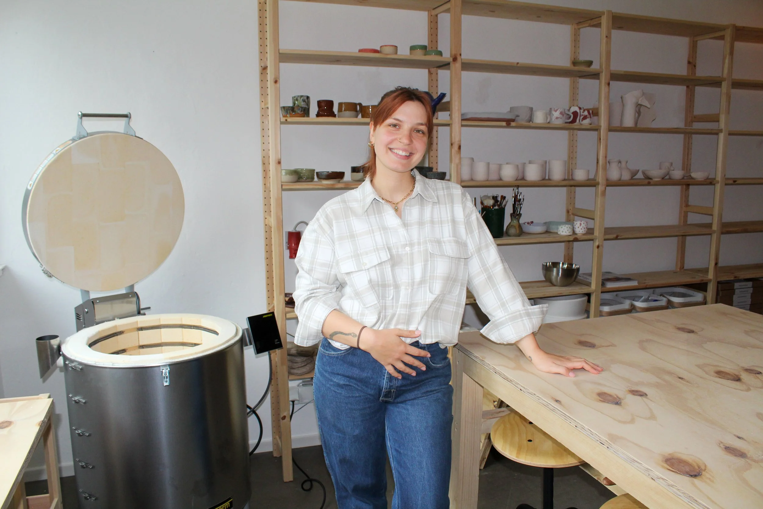 Gründerin Rebekka, eine junge, lächelnde Frau in Jeans und Flanellhemd, steht in ihrem Töpferstudio neben einem Holztisch, im Hintergrund offene Holzregale mit Keramik.