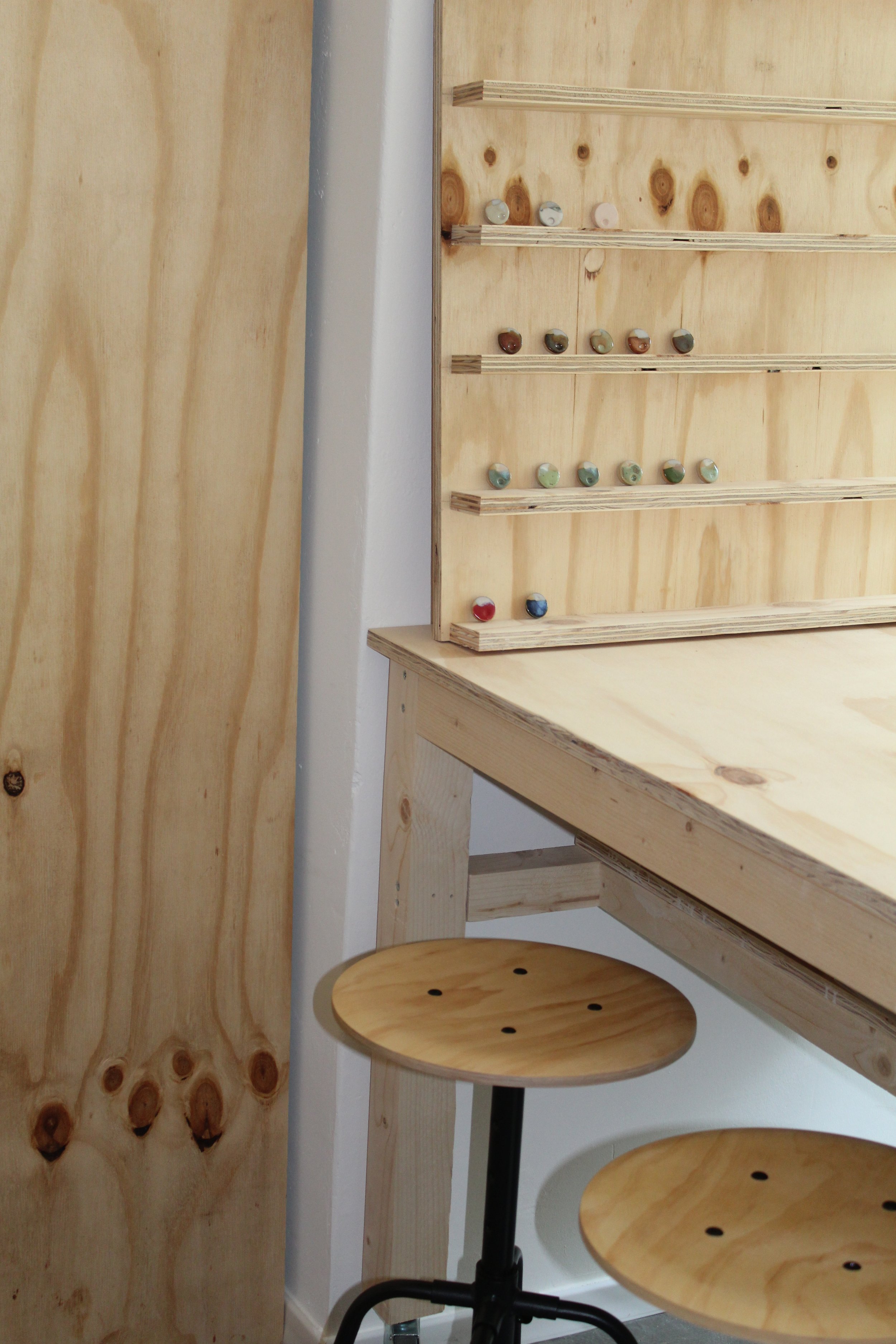 Holzregal mit Glasur-Beispielen, daneben ein hölzerner Drehstuhl an einem Werktisch.
