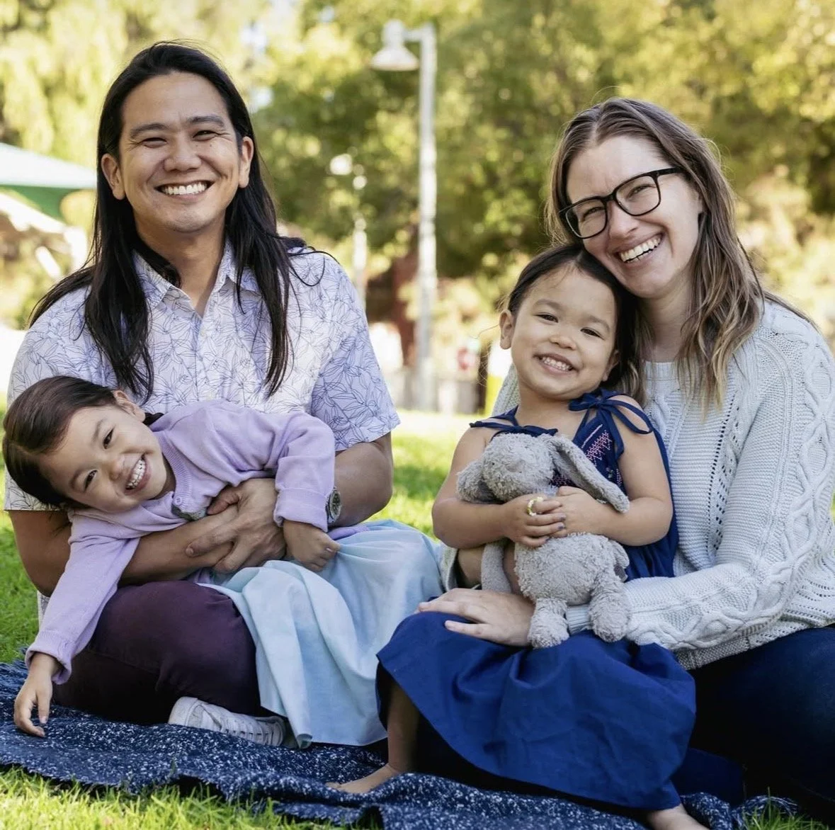 Four people sitting outdoors on a grassy area, smiling and enjoying the day. Two women and two young girls, one girl holding a stuffed animal.