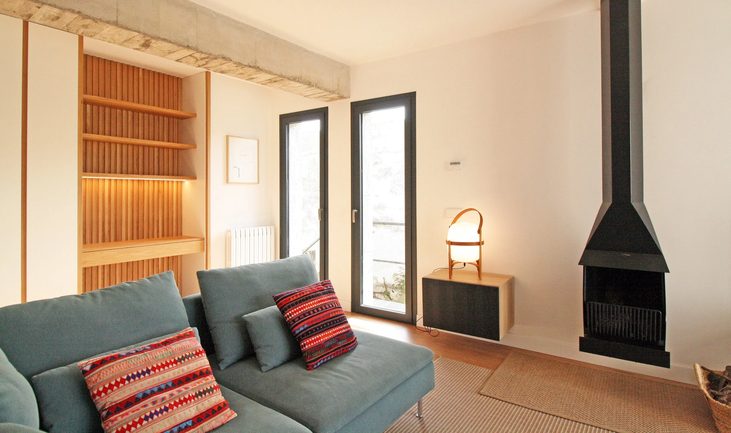 Sala moderna con sofá azul, cojines decorativos con patrones coloridos, paredes blancas, ventanas grandes, lámpara de mesa de madera, chimenea negra y una alfombra de fibras naturales.