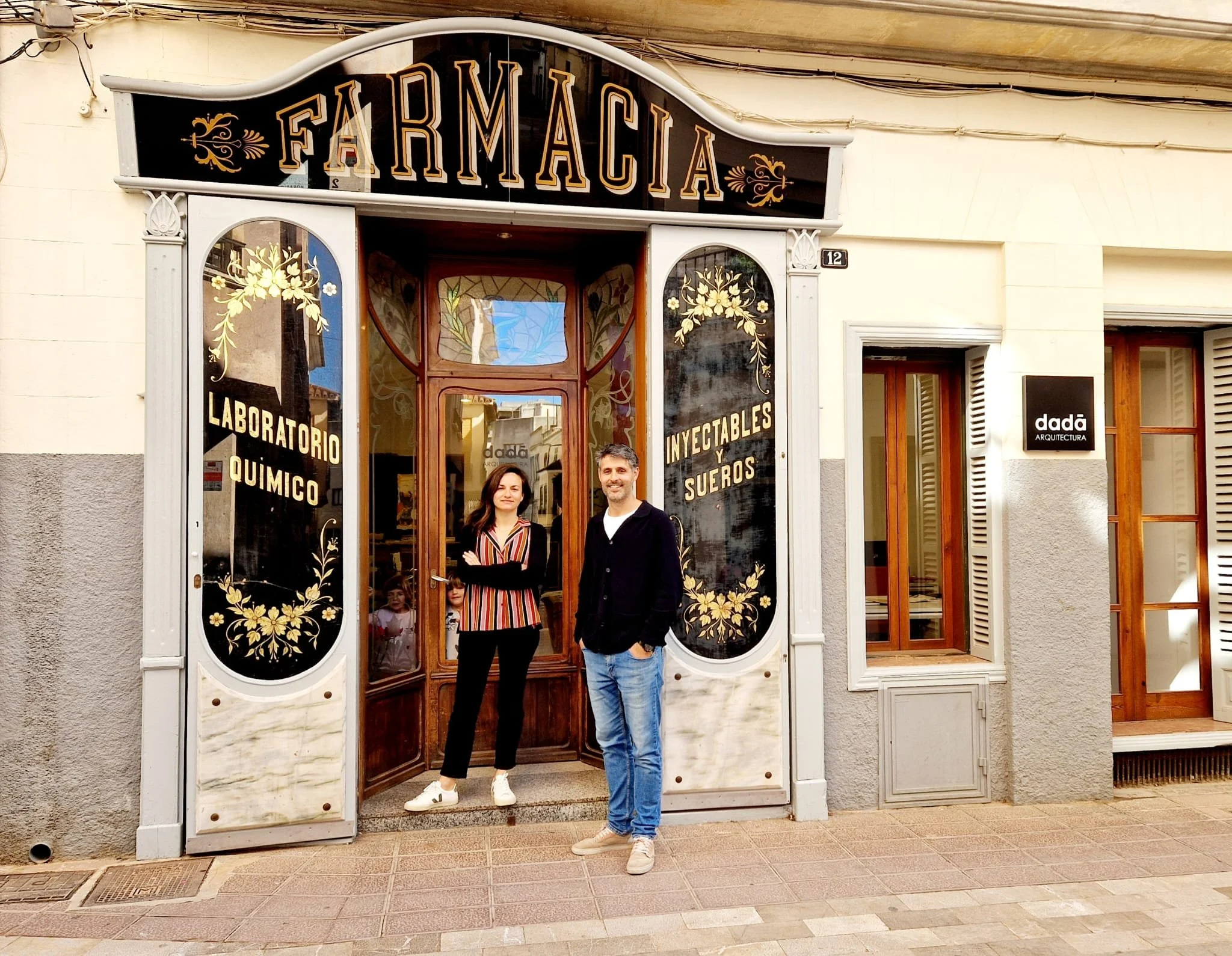 Fachada de una farmacia con señalización en letras grandes y ornamentación decorativa negra con dorado, ventanas con diseños florales, y dos personas jóvenes sonriendo delante de la entrada.