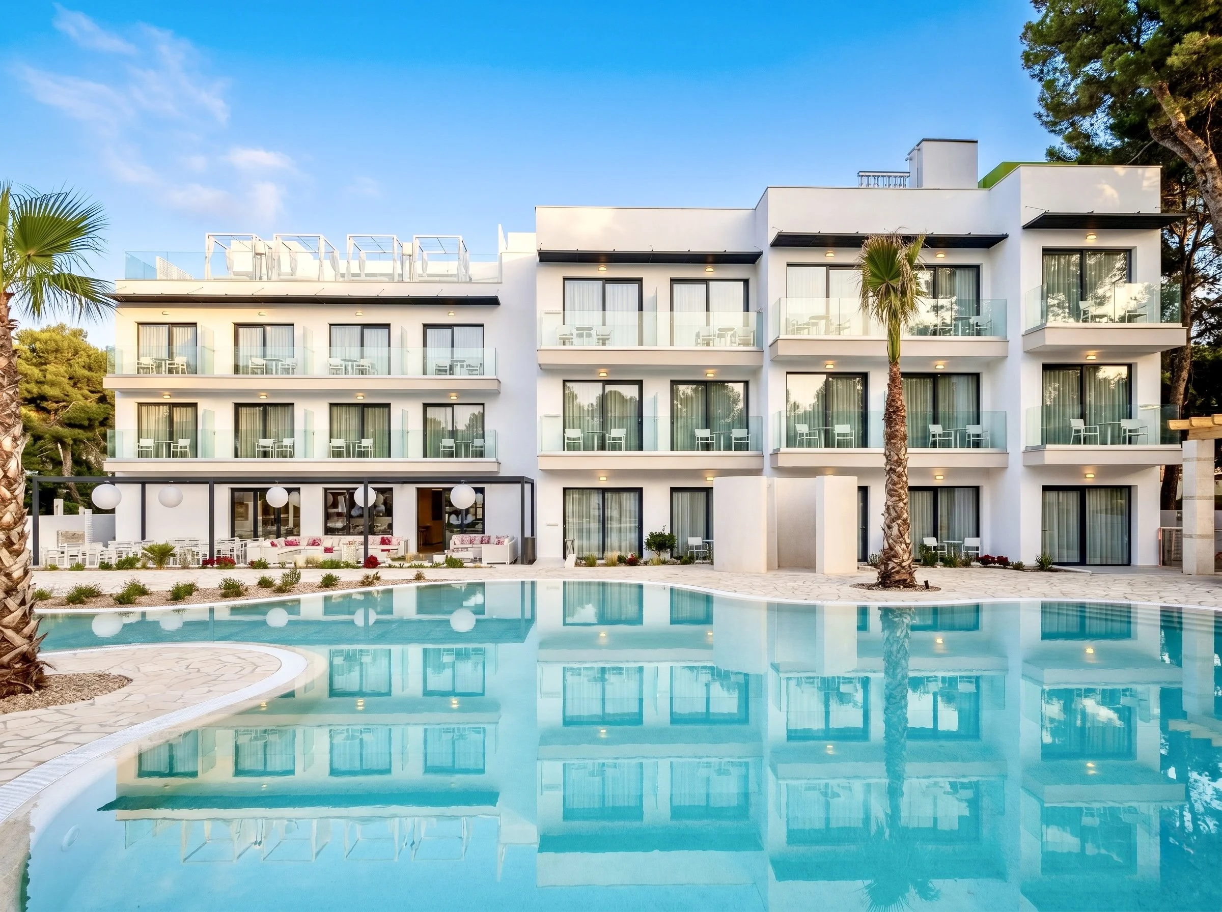 Hotel moderno con varias terrazas y balcones de cristal, junto a una piscina con agua clara rodeada de palmeras y áreas verdes, en un día despejado.