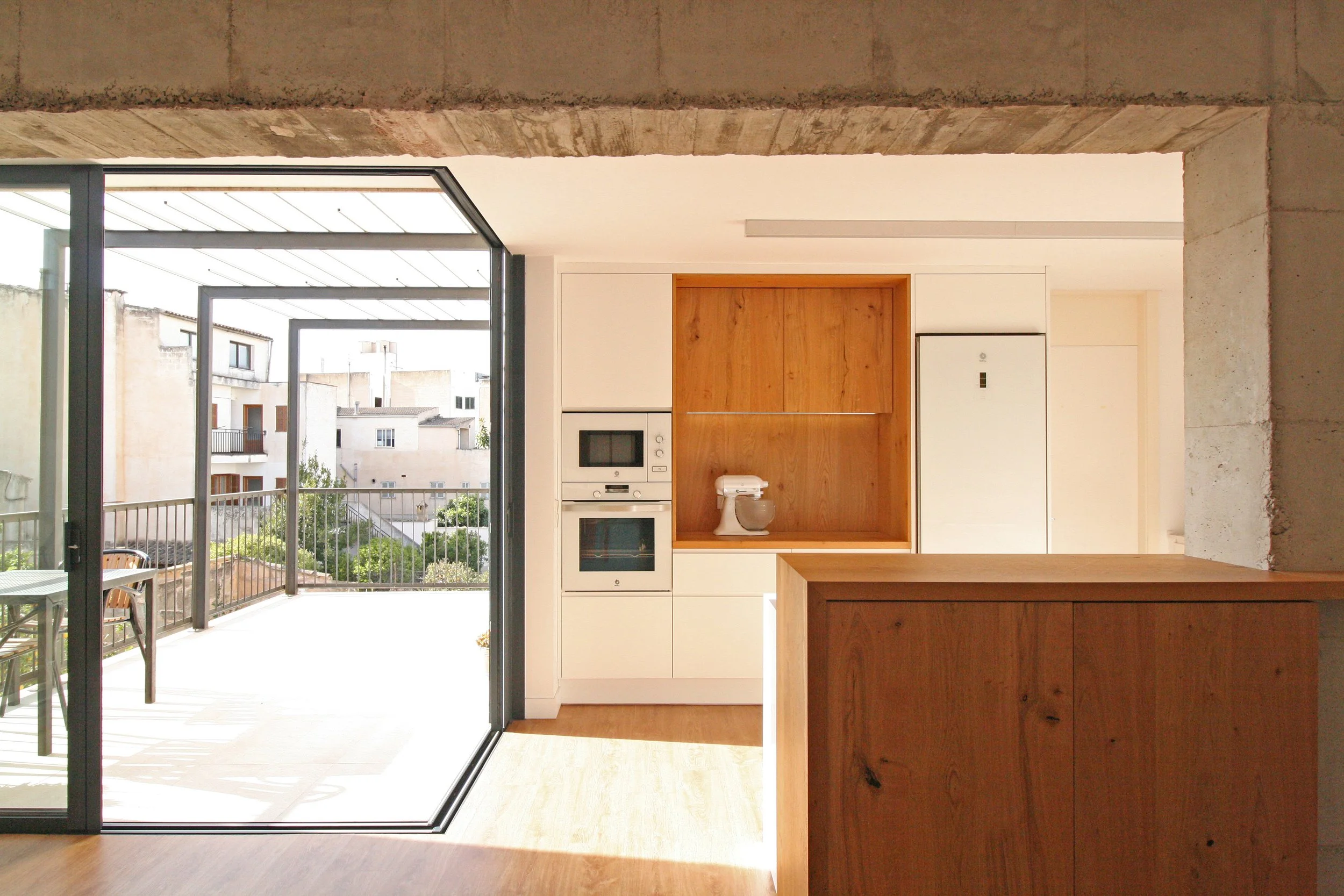 interior de un apartamento con balcón, vistas de edificios exteriores, cocina moderna con electrodomésticos y muebles de madera, paredes blancas y suelo de madera
