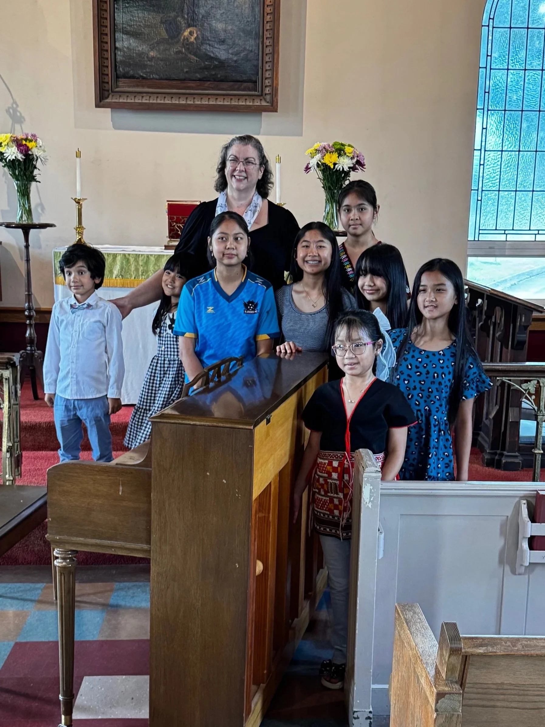 A group of children and a woman standing inside All Saints' behind a piano, with two flower arrangements and candles on the altar behind them, and a stained glass window on the right.