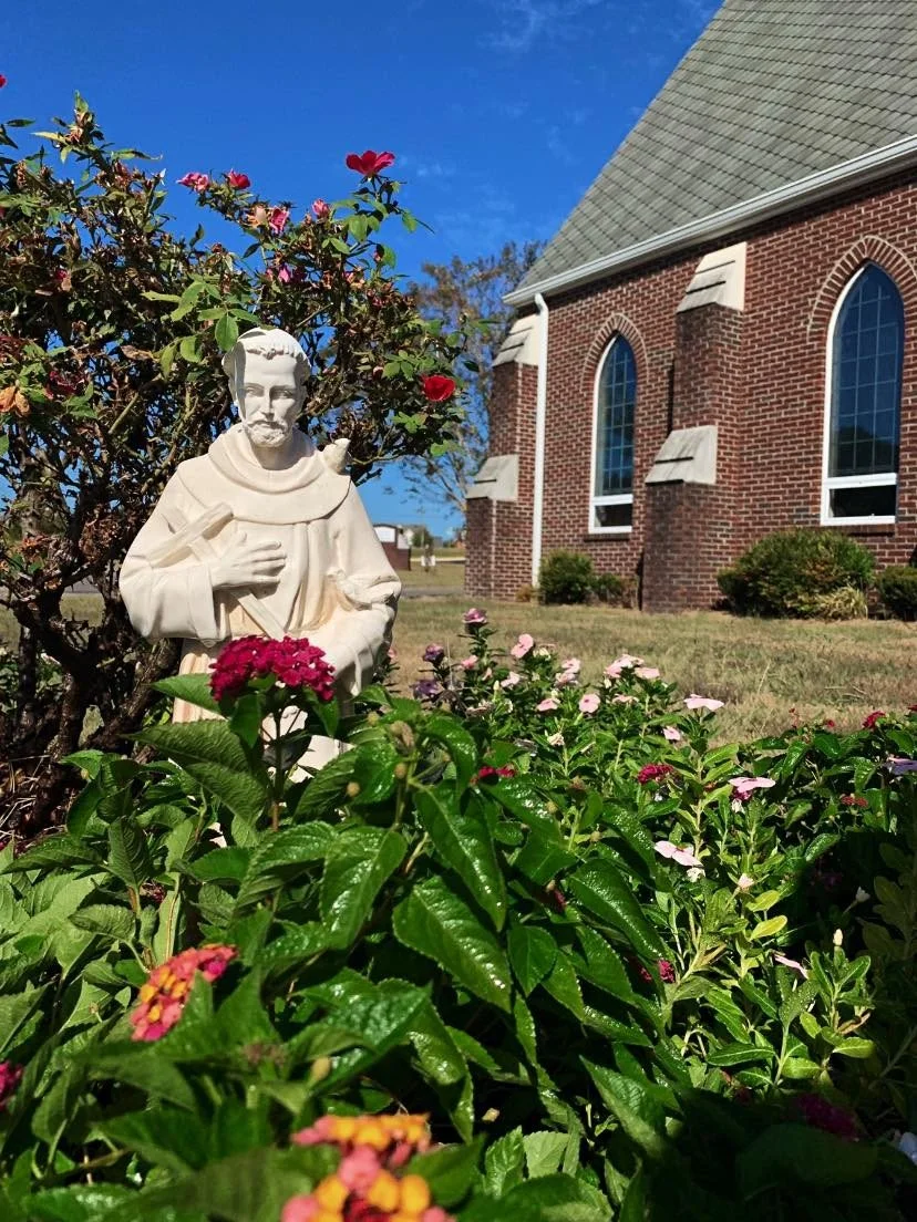 A white statue of St. Francis of Assisi surrounded by pink and red flowers in front of All Saints' with arched windows and a green roof. The sky is blue with some clouds.