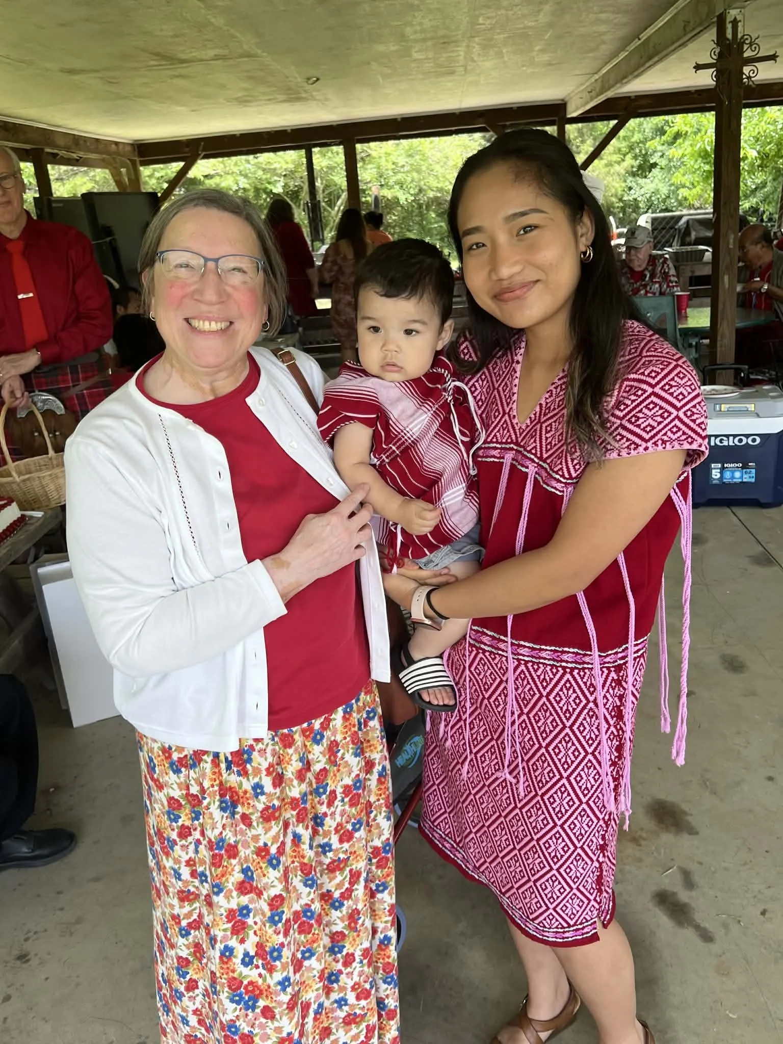 Three women and a child standing together indoors, smiling, with others in the background at a gathering or celebration.