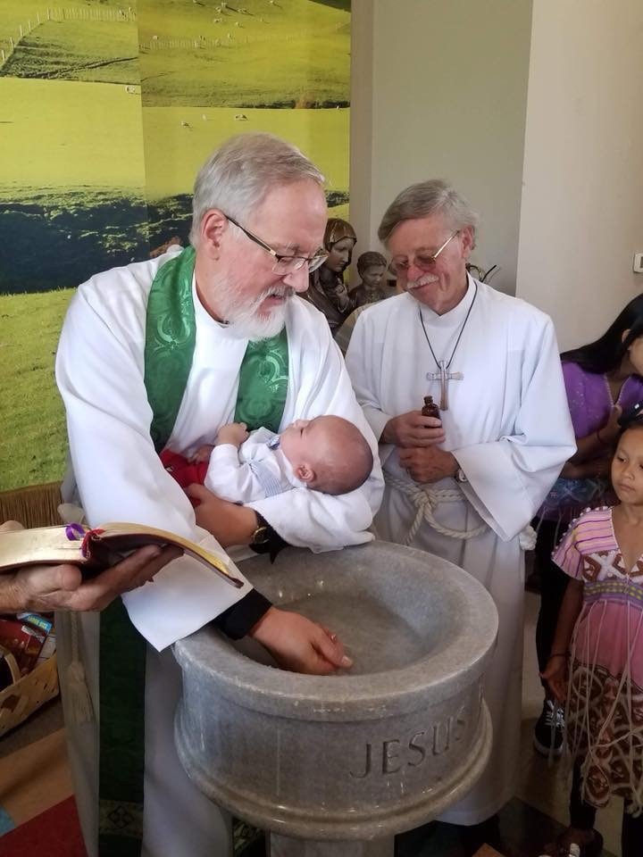 A baptism ceremony with a baby being baptized by Vicar Robert Rhea at All Saints'. The priest holds the baby over a stone basin labeled 'JESUS' while another clergy member stands nearby. A person holds a book and an oil bottle.