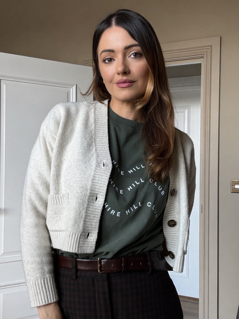 Young woman with long brown hair, wearing a white cardigan over a dark green T-shirt, standing in a room with white doors and beige walls.
