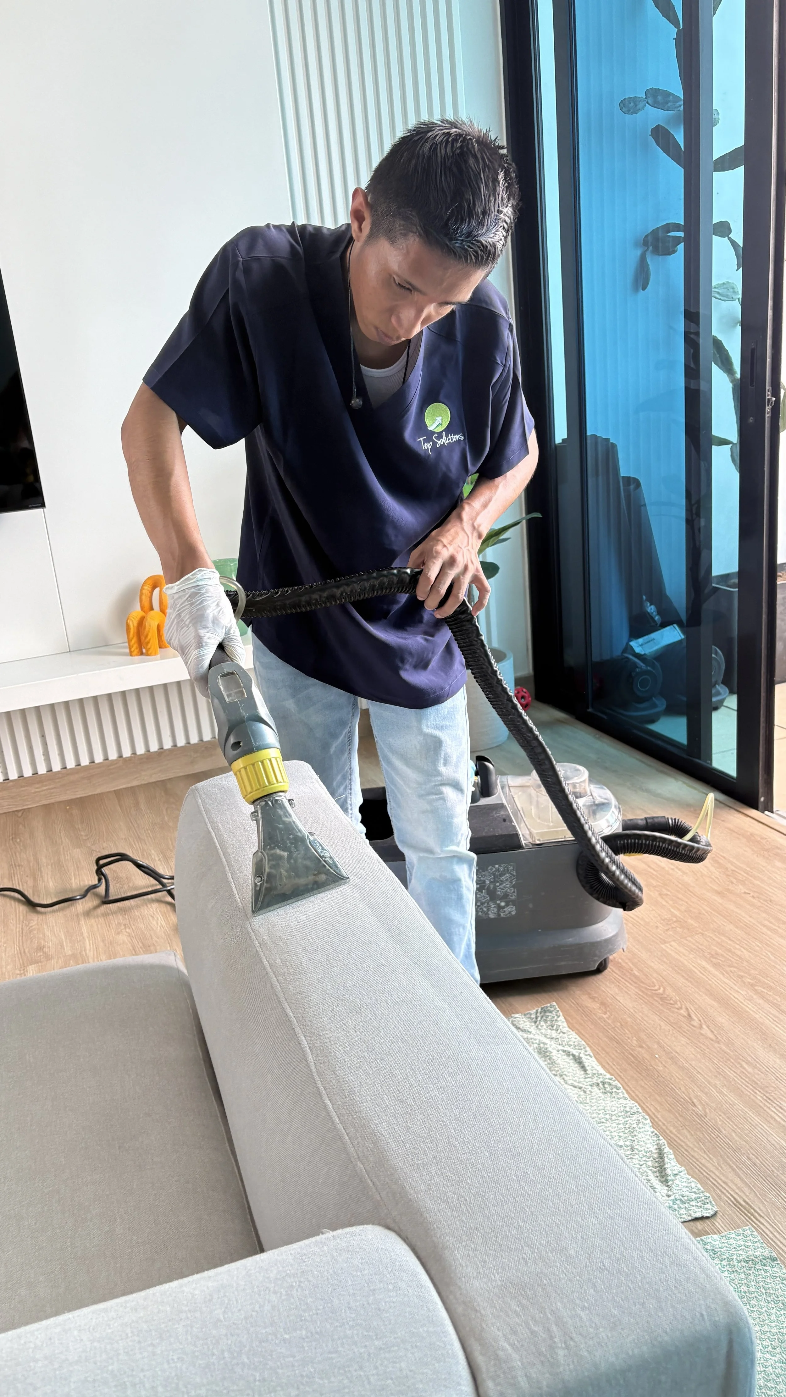 Hombre limpiando el sofá con una aspiradora en una sala moderna con paredes de vidrio y plantas en el fondo.