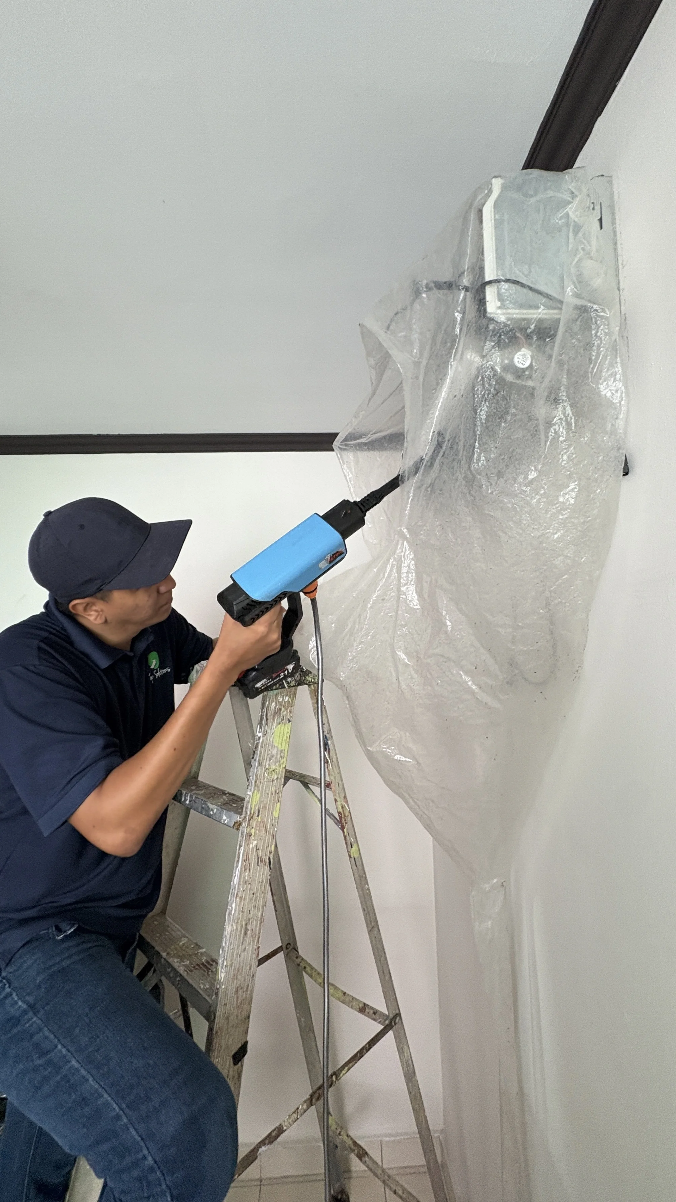 Persona usando una pistola de calor para quitar pegamento o plástico de una pared, de pie en una escalera de aluminio.