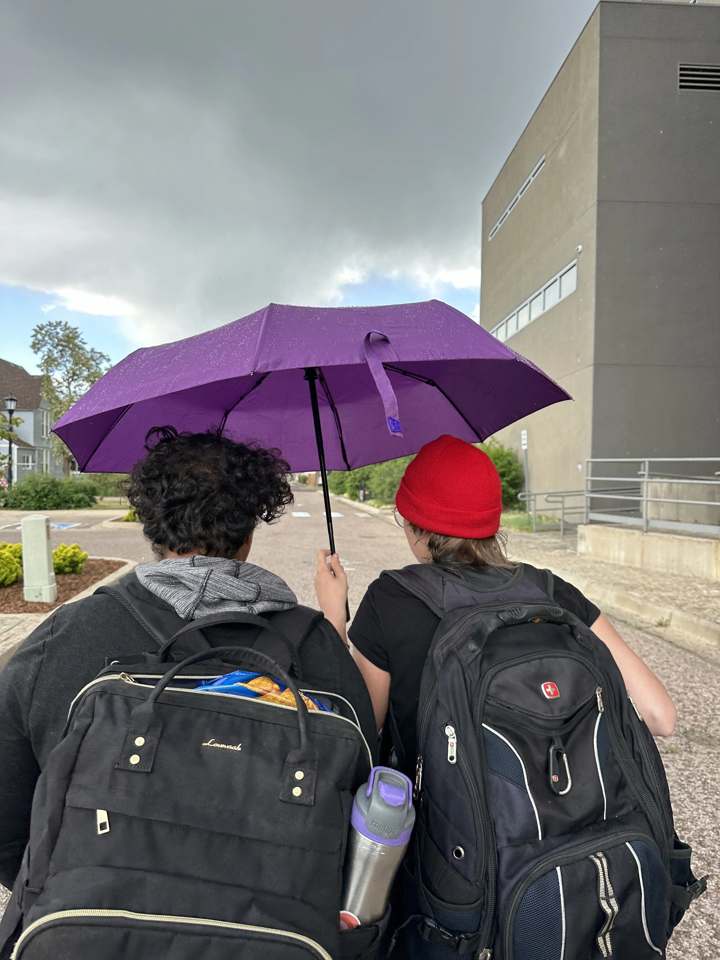 Two people with backpacks, one holding a purple umbrella, standing outdoors on a cloudy day in front of a modern building.