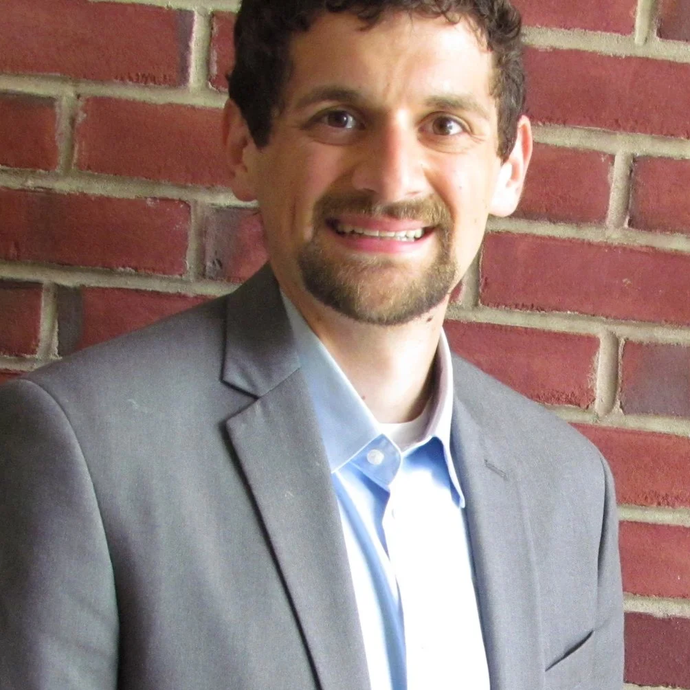 A man in a gray suit smiling in front of a red brick wall.