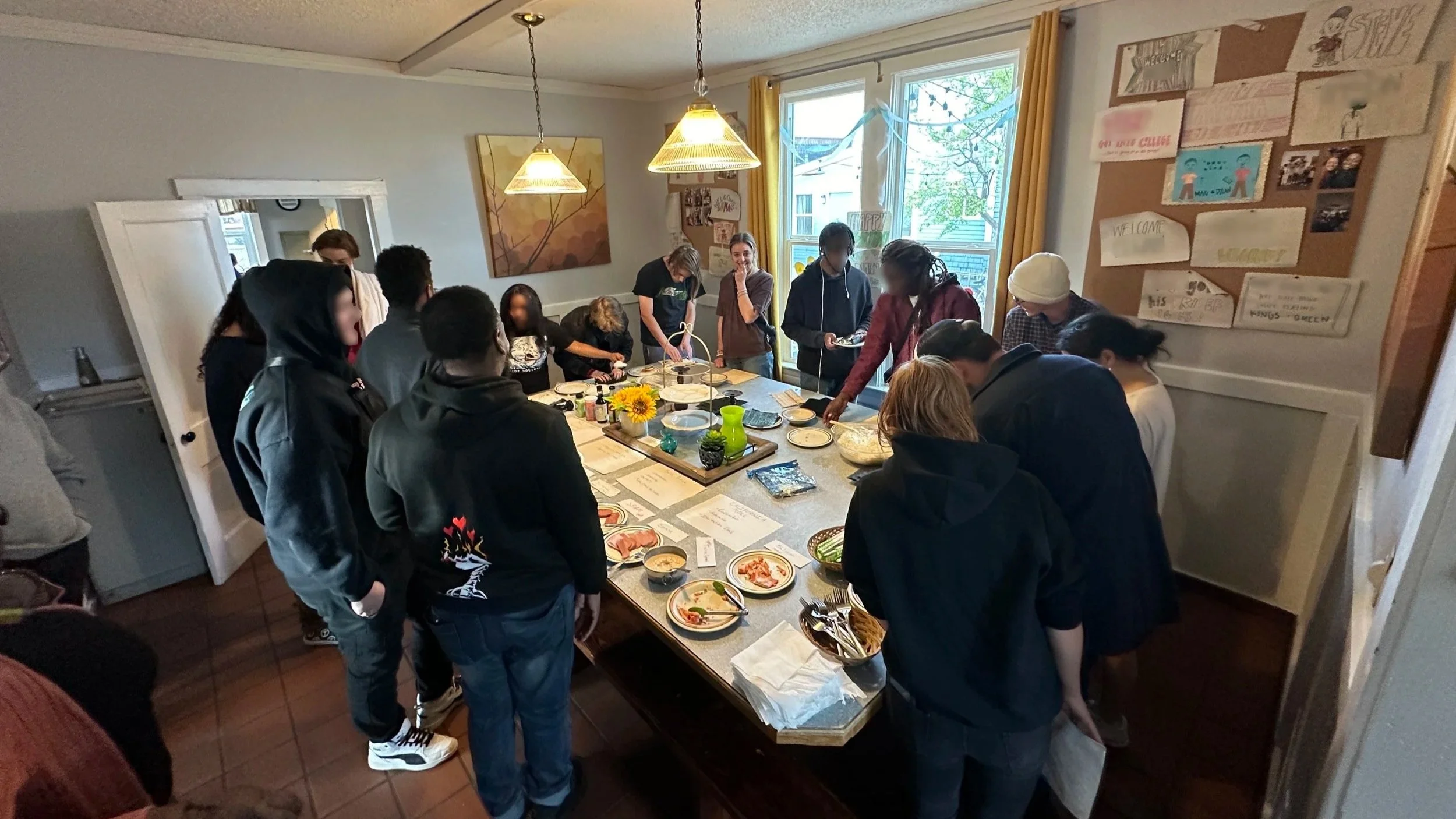 Group of people gathered around a dining table with food and plates in a cozy kitchen or dining room.