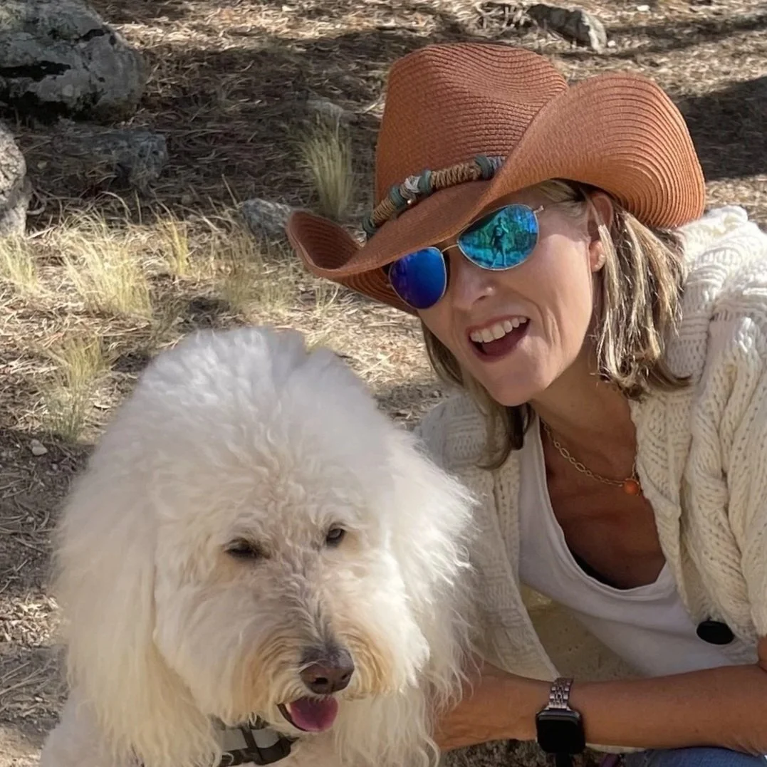 A woman wearing a large hat, sunglasses, and a white sweater, smiling and taking a selfie with a fluffy white dog outdoors in a desert-like environment.