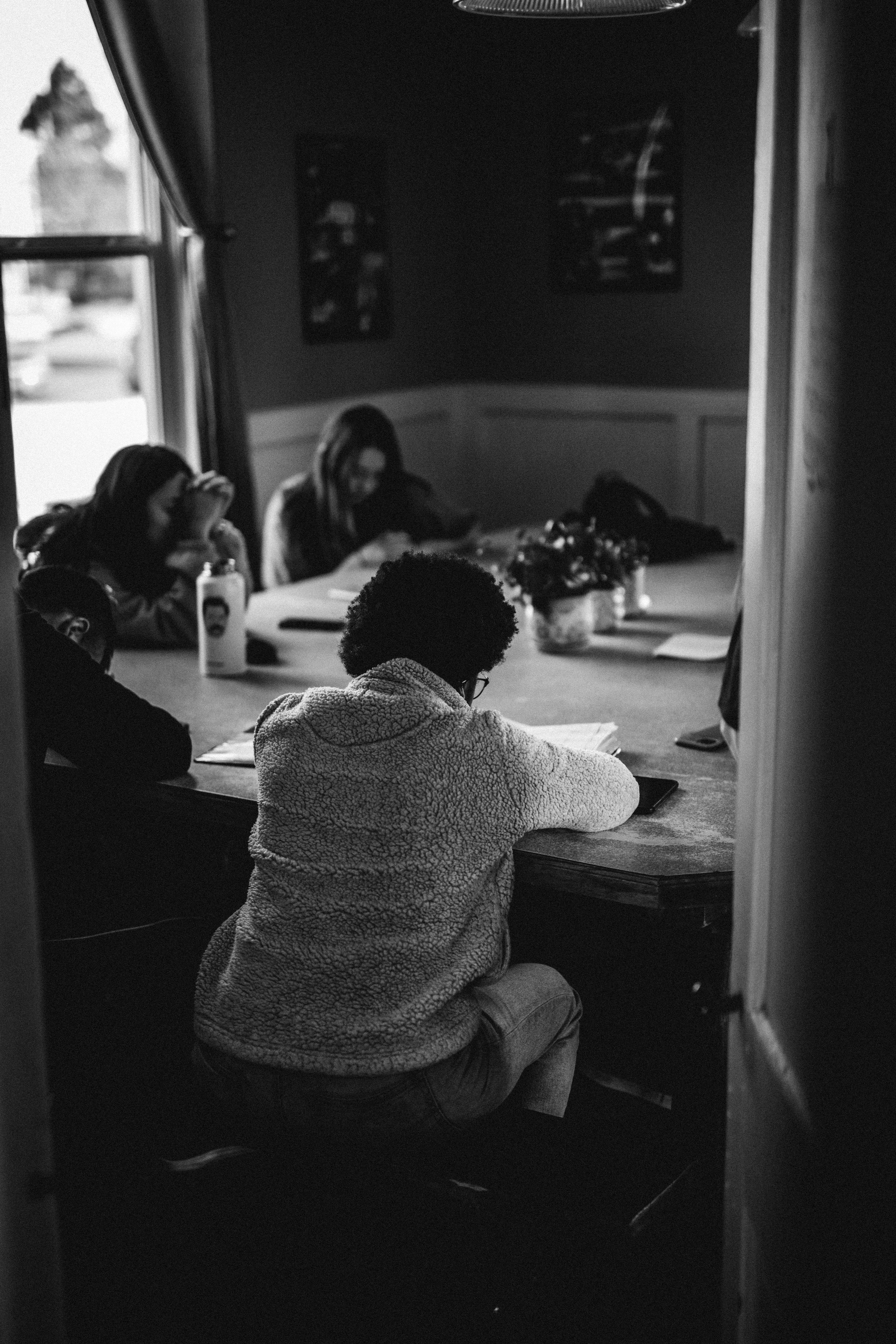 A group of children sitting at a table in a room, engaged in an activity like studying or reading, with a window and some picture frames on the wall in the background.
