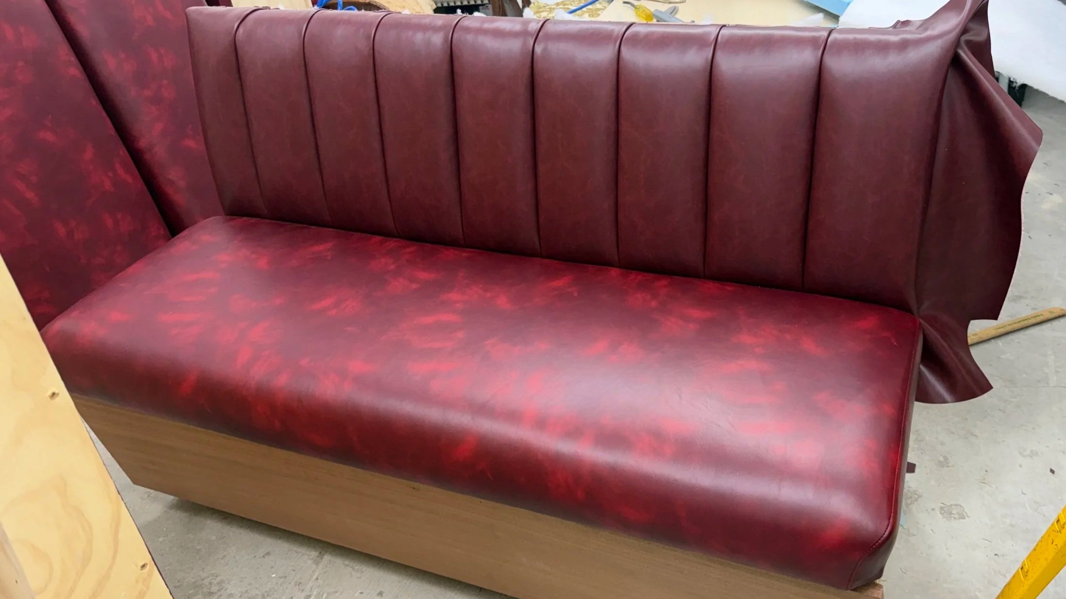 A red booth-style upholstered seat with a high backrest and vertical stitching, resting on a wooden base, placed in a warehouse or workshop setting.