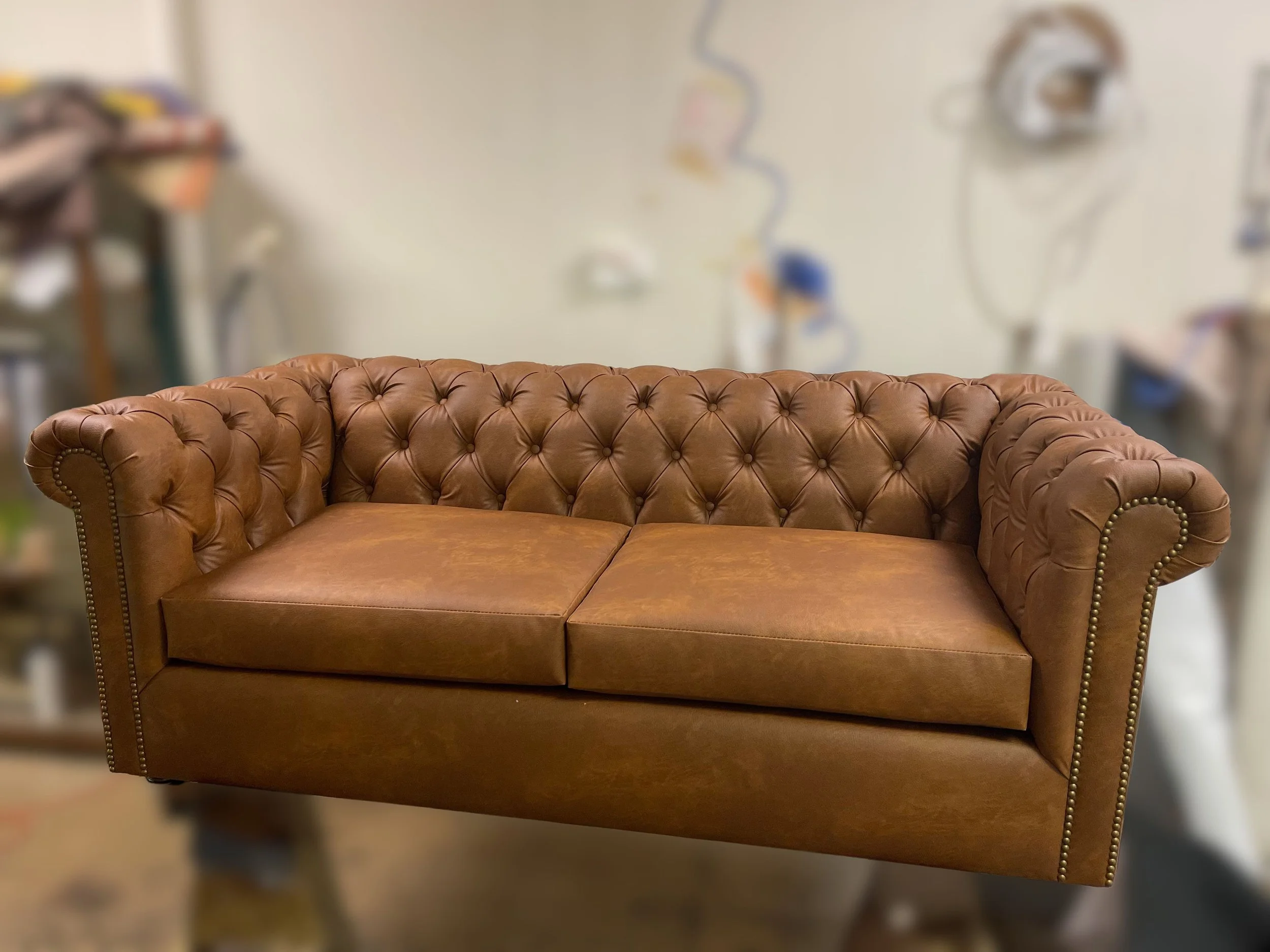 A brown leather tufted sofa with rolled arms and brass nailhead trim, situated in a room with a blurred background of medical equipment and supplies.