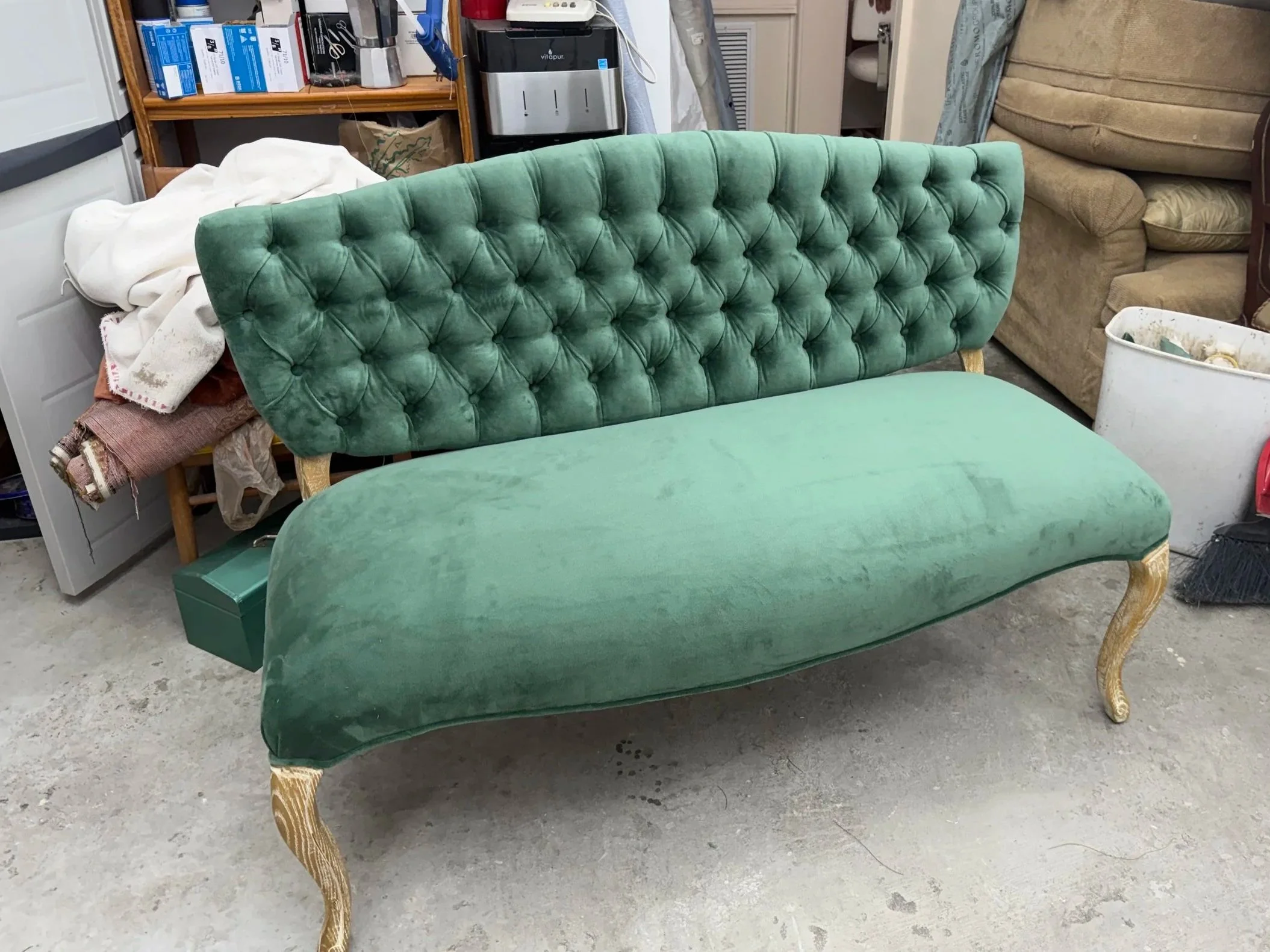 A vintage green velvet tufted chaise lounge with ornate gold legs in a cluttered storage room, with various furniture and household items in the background.