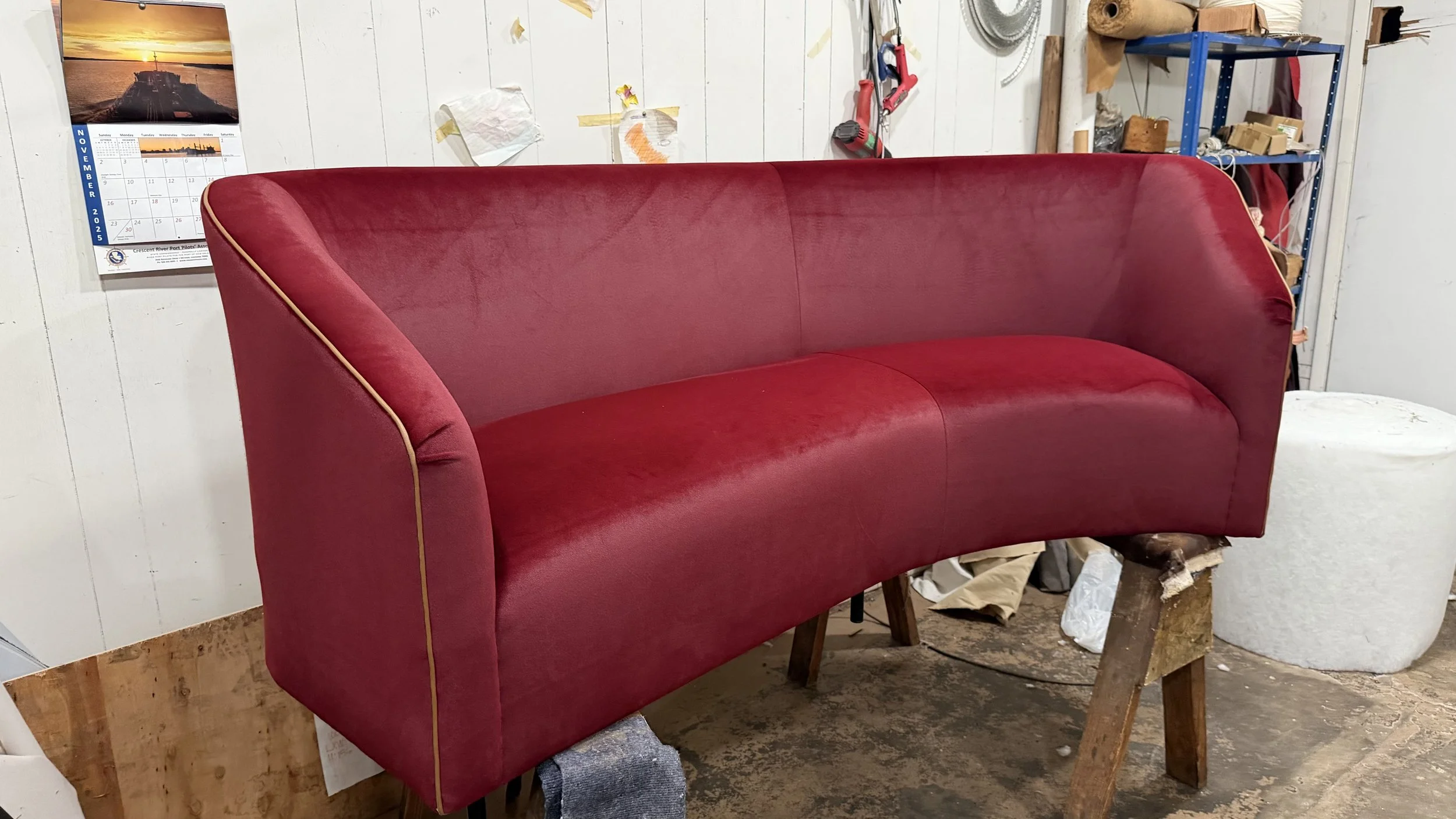 A red upholstered sofa with gold piping in a workshop setting, surrounded by tools, a calendar, and various supplies.
