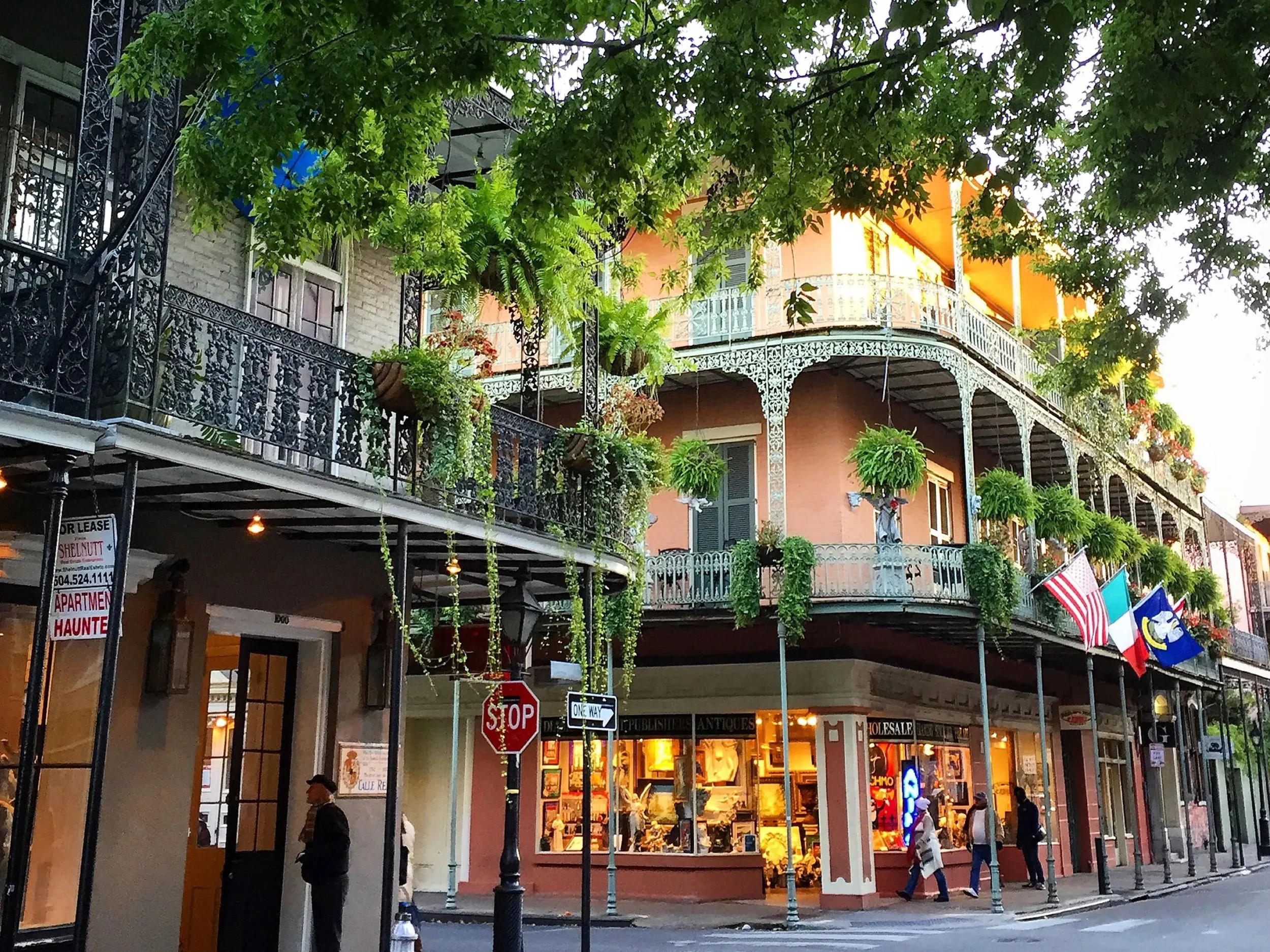 Street scene with retail shops, several American flags, street signs, and a building with decorative iron balconies and plants, partly shaded by green leafy trees.