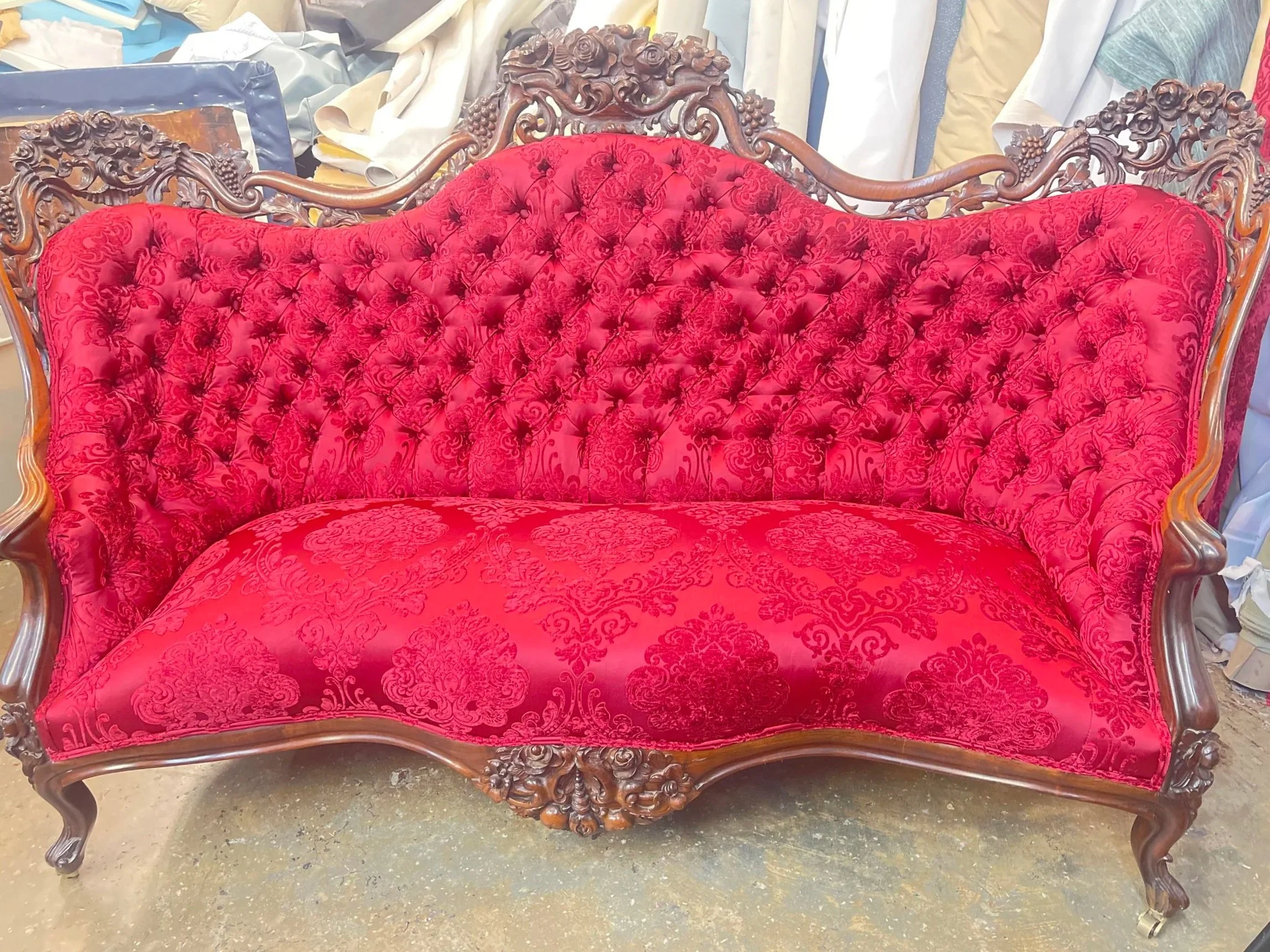 An ornate vintage sofa with a carved wooden frame and red tufted upholstery with intricate patterns, situated in what appears to be a storage or workshop area.