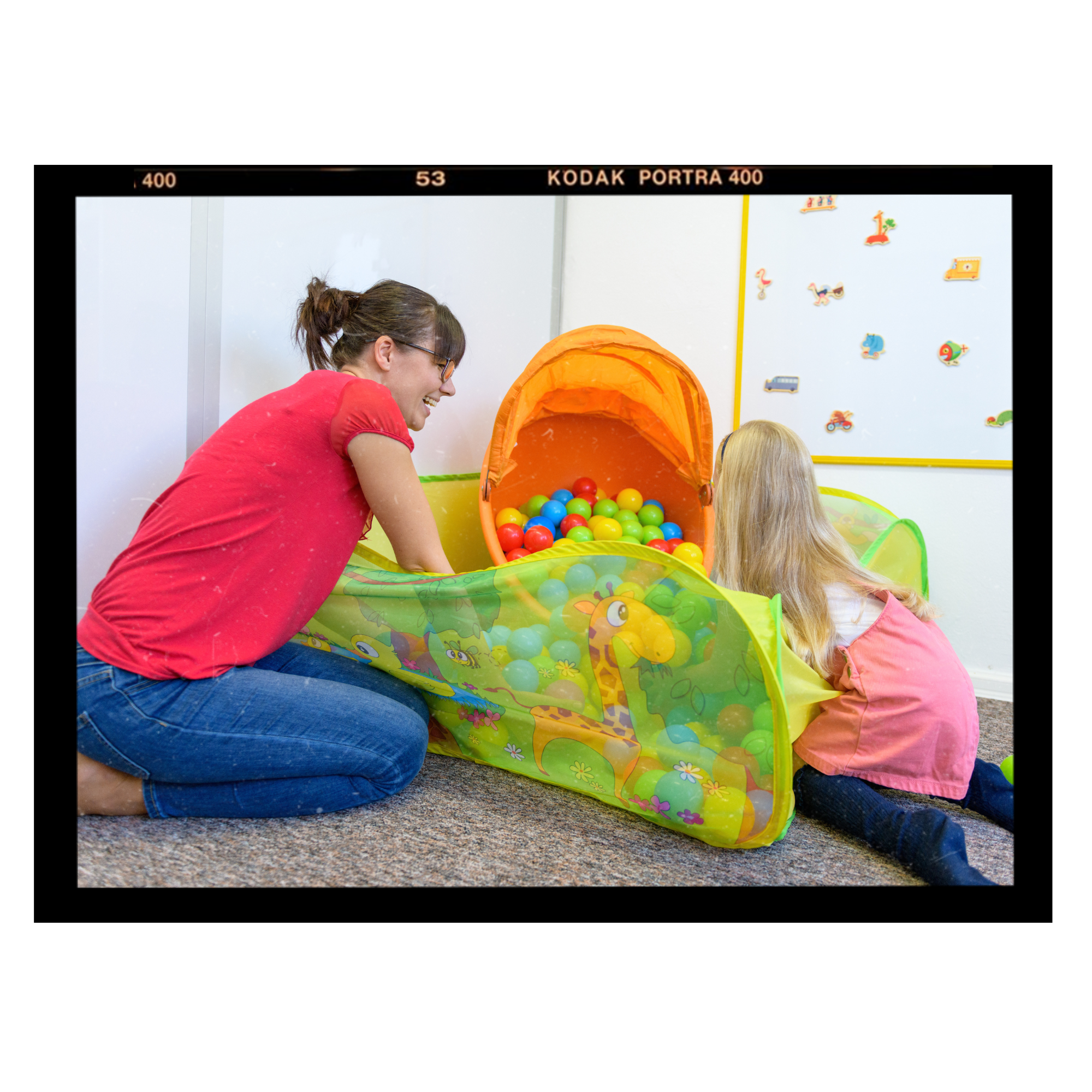 A woman and a young girl are playing with a colorful ball pit that has a yellow and green design on the outside. The woman is wearing a red shirt and blue jeans, and the girl is wearing a pink shirt. They are inside a room with white walls decorated with animal stickers and a yellow dividing line, and a whiteboard is visible in the background.