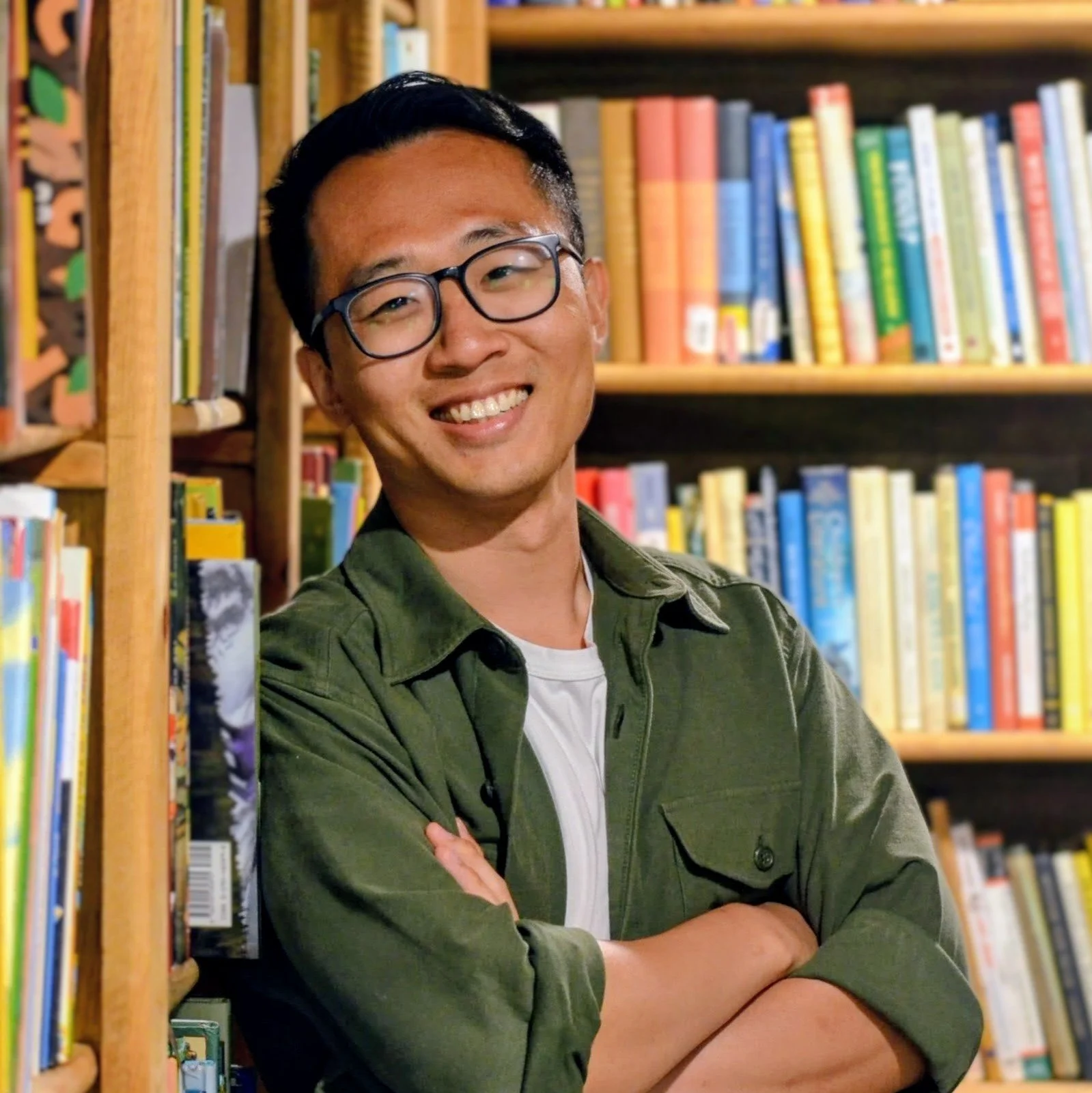 A photo of the author, Roger Lam: a young Chinese American man with black hair and glasses standing in front of a colorful bookshelf.