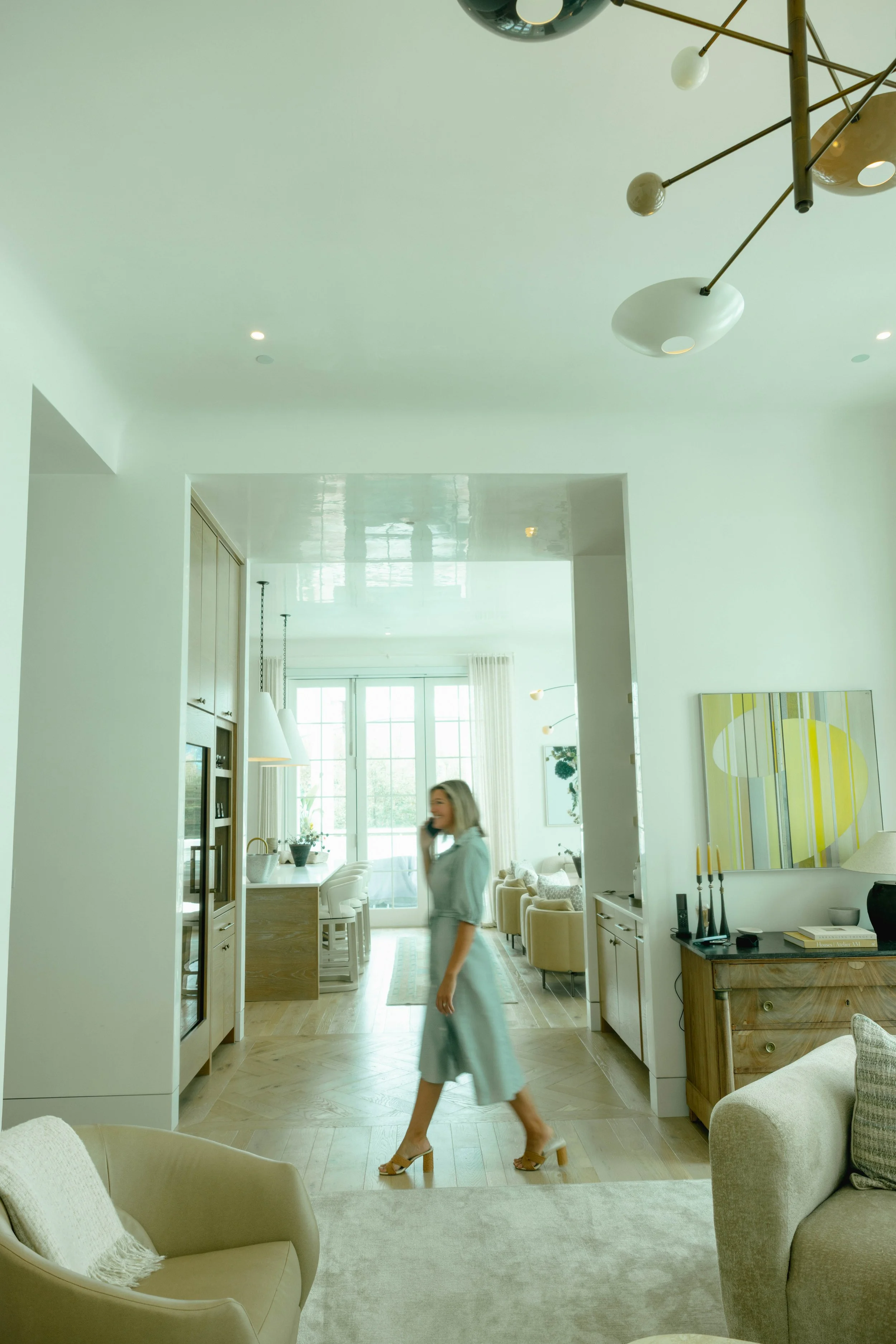 Modern living room with beige and white furniture, a light hardwood floor, large windows letting in natural light, and a woman with blonde hair in a light green dress walking across the room.